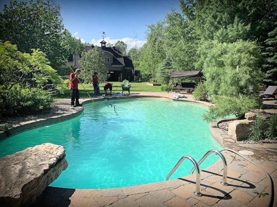Trois personnes nettoient une piscine turquoise éclatante entourée de dalles de pierre, d'arbres luxuriants et d'une maison en arrière-plan.