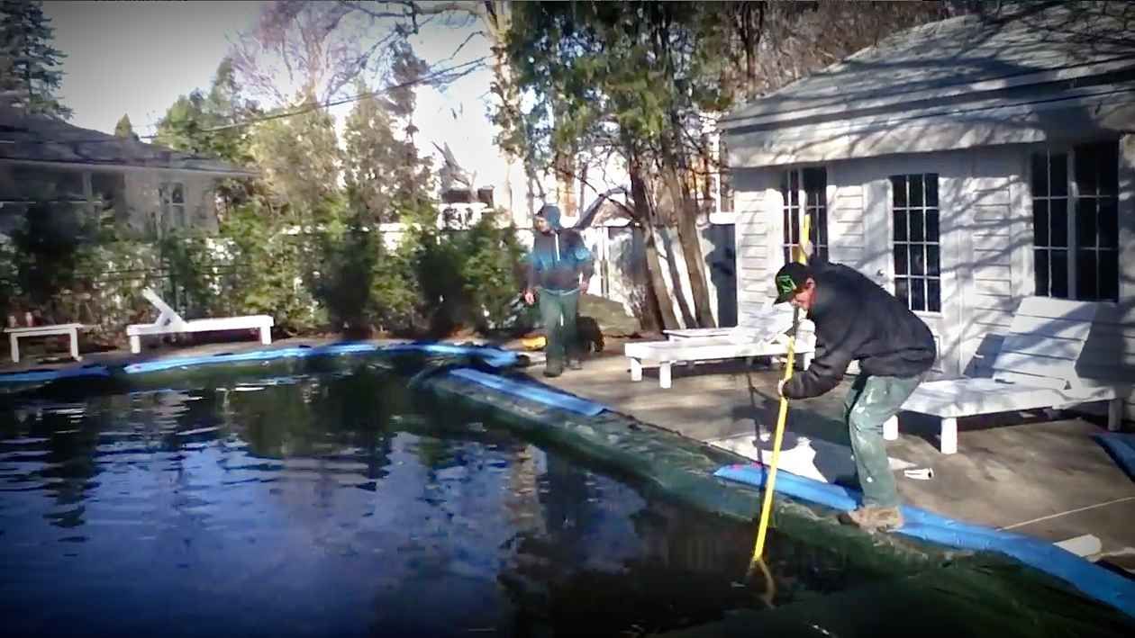 Une personne utilise un outil à long manche pour nettoyer une piscine dans un jardin, à côté d'un petit bâtiment blanc et de chaises de terrasse.