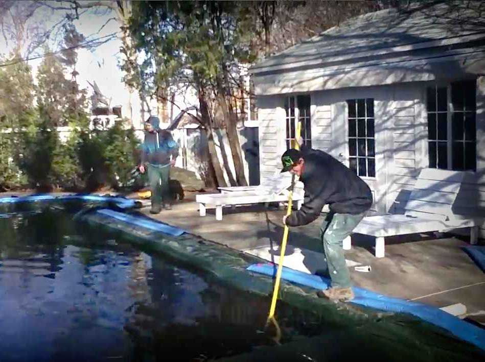 Une personne écume les feuilles mortes d'une piscine située dans un jardin près d'une maison blanche et d'une terrasse.