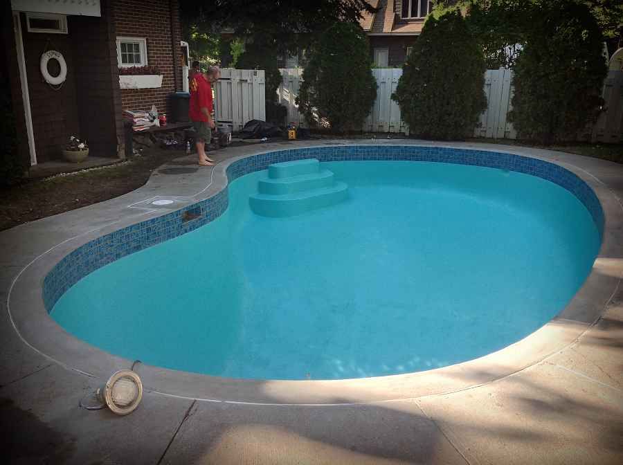 Piscine de jardin avec eau bleu vif et terrasse, à côté d'une maison et d'une clôture.