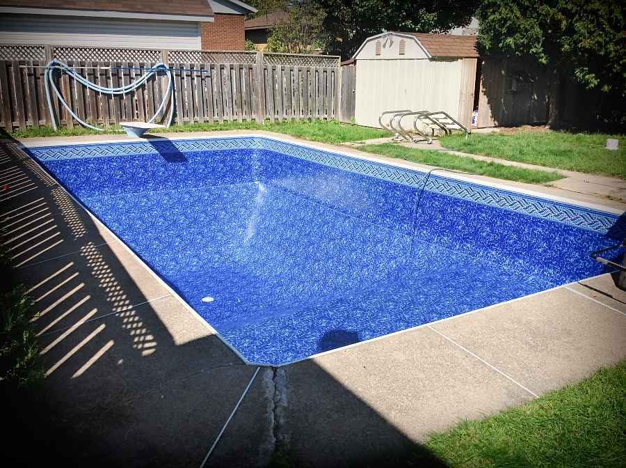 Piscine de jardin recouverte d'une bâche bleue, avec clôture, pelouse et abri de jardin en arrière-plan.