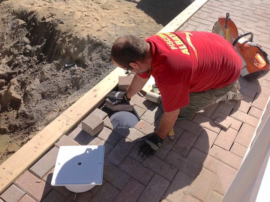 Une personne vêtue d'une chemise rouge est agenouillée sur une allée en briques, en train d'installer un appareil sanitaire près d'une tranchée ouverte.