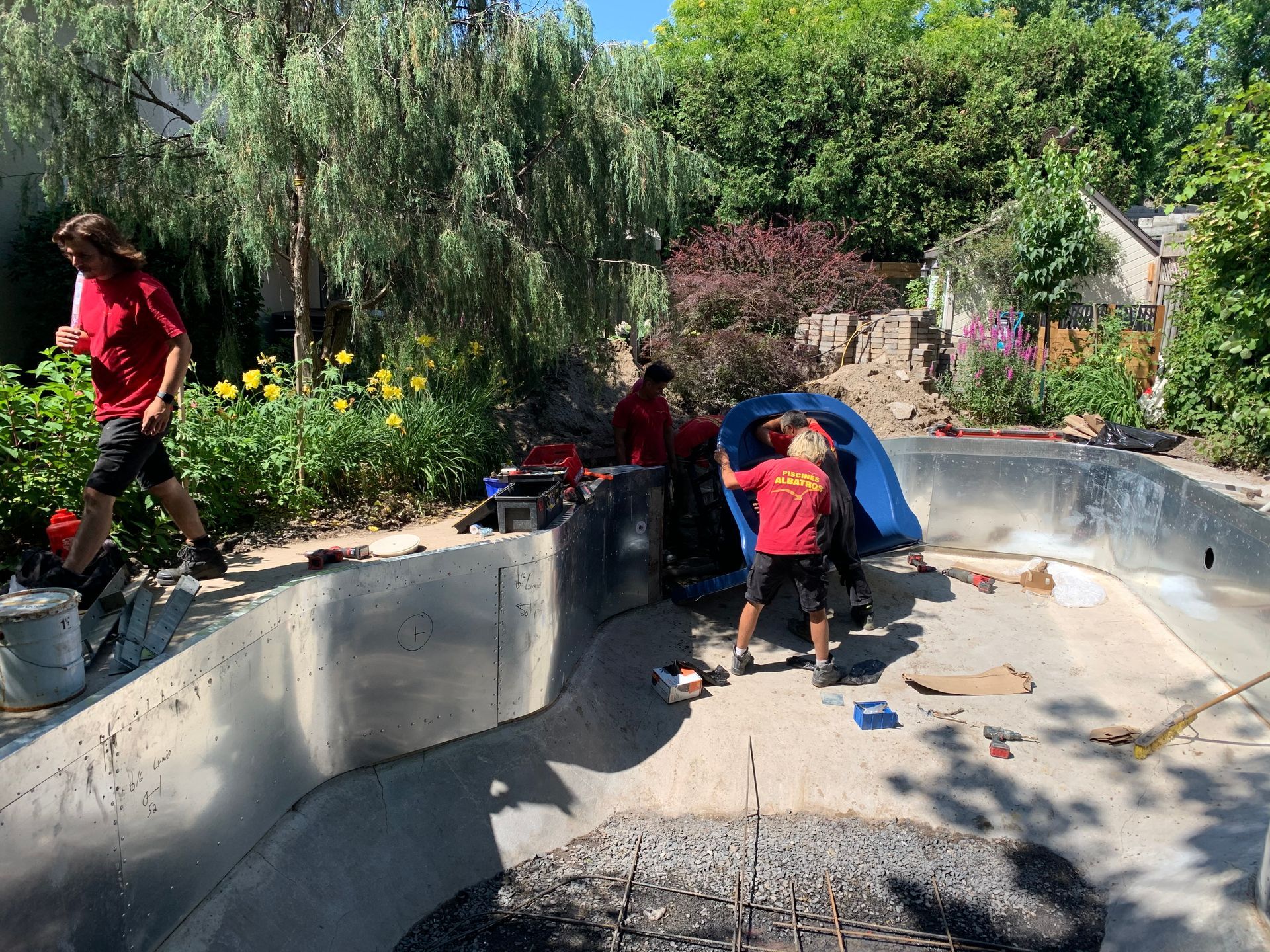 Des personnes vêtues de chemises rouges travaillent à la construction ou à la réparation des parois métalliques d'une piscine extérieure dans un jardin ensoleillé.