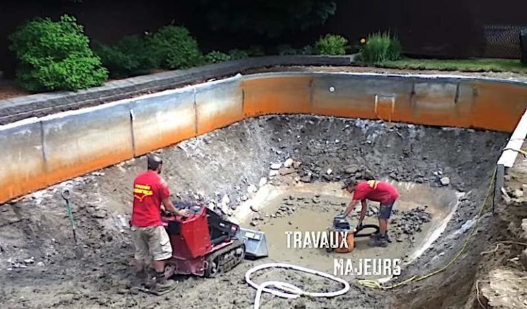 Deux ouvriers en chemises rouges manœuvrent des machines à l'intérieur d'une piscine vide et boueuse, en cours de fouilles ou de rénovation.