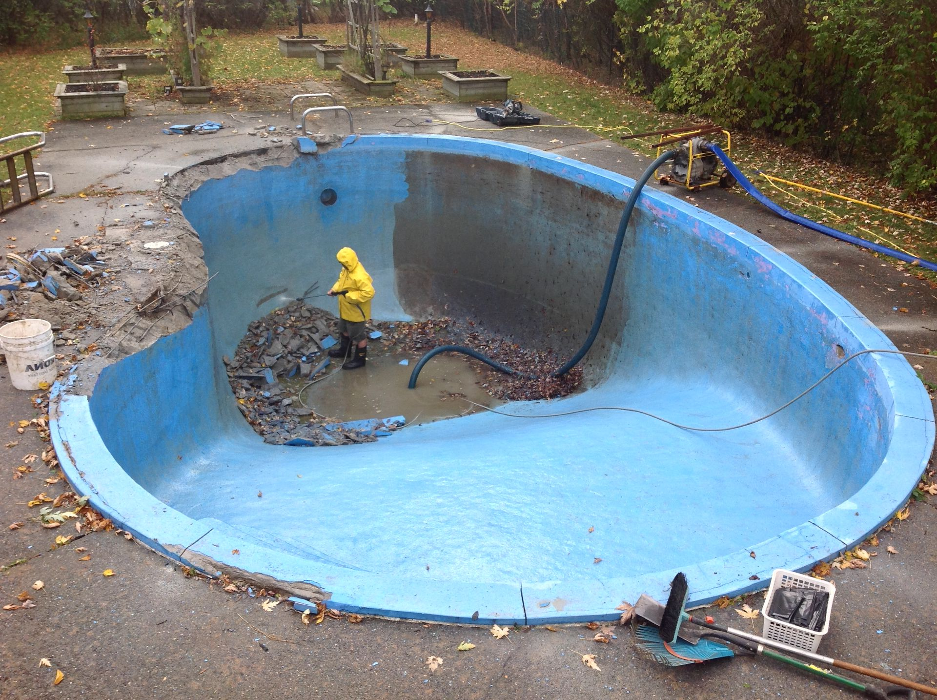 Un ouvrier en imperméable jaune nettoie une piscine bleue vidée à l'aide d'un tuyau d'arrosage.
