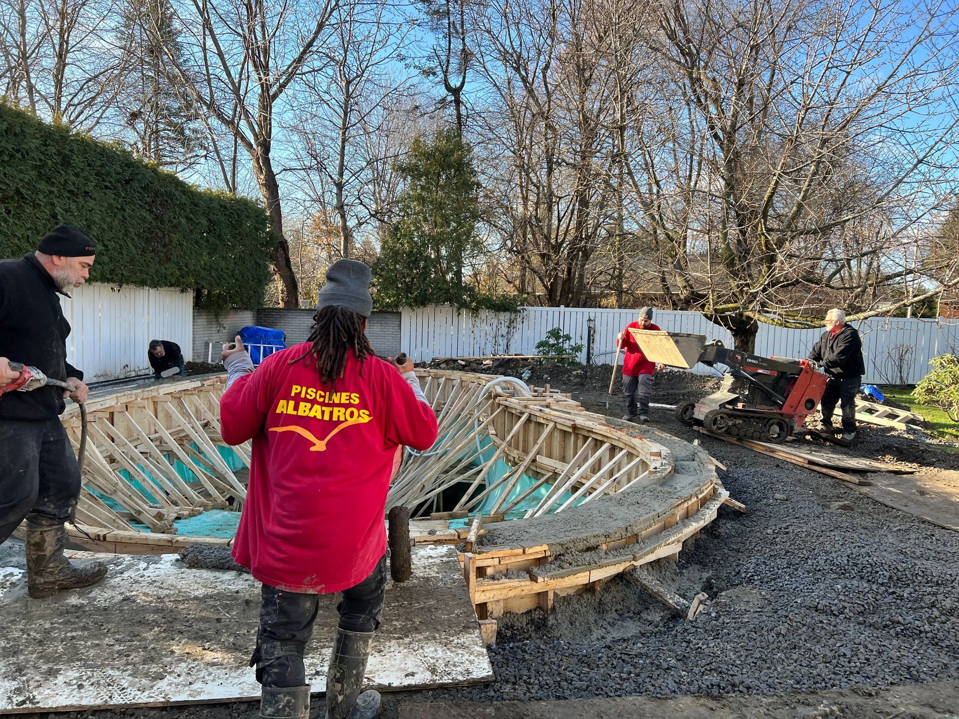 Construction workers build a curved concrete pool edge in a backyard with wooden framing and surrounding gravel.