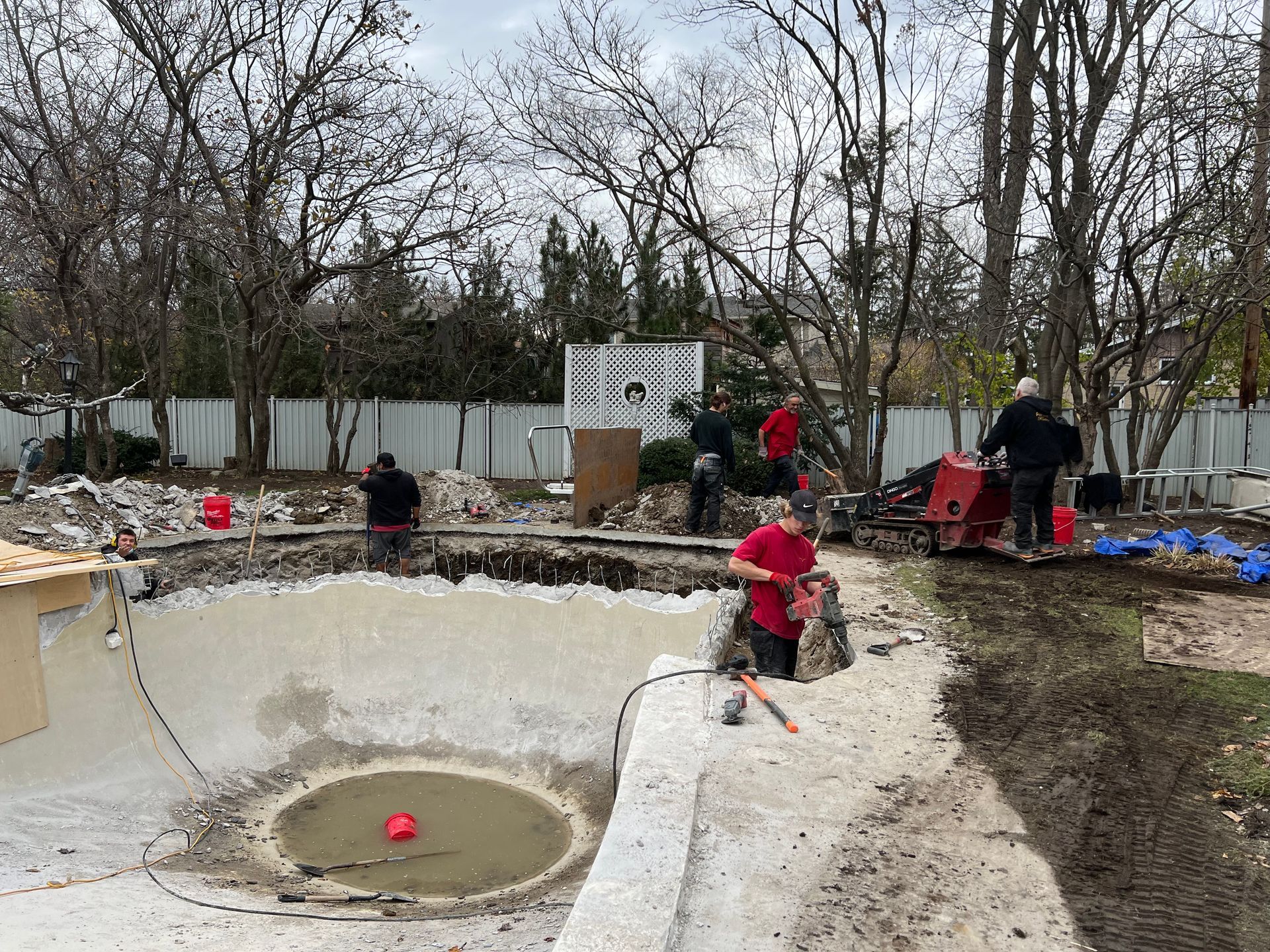 Des ouvriers réparent une piscine vide dans un jardin boisé, avec des outils et des matériaux autour du bassin.