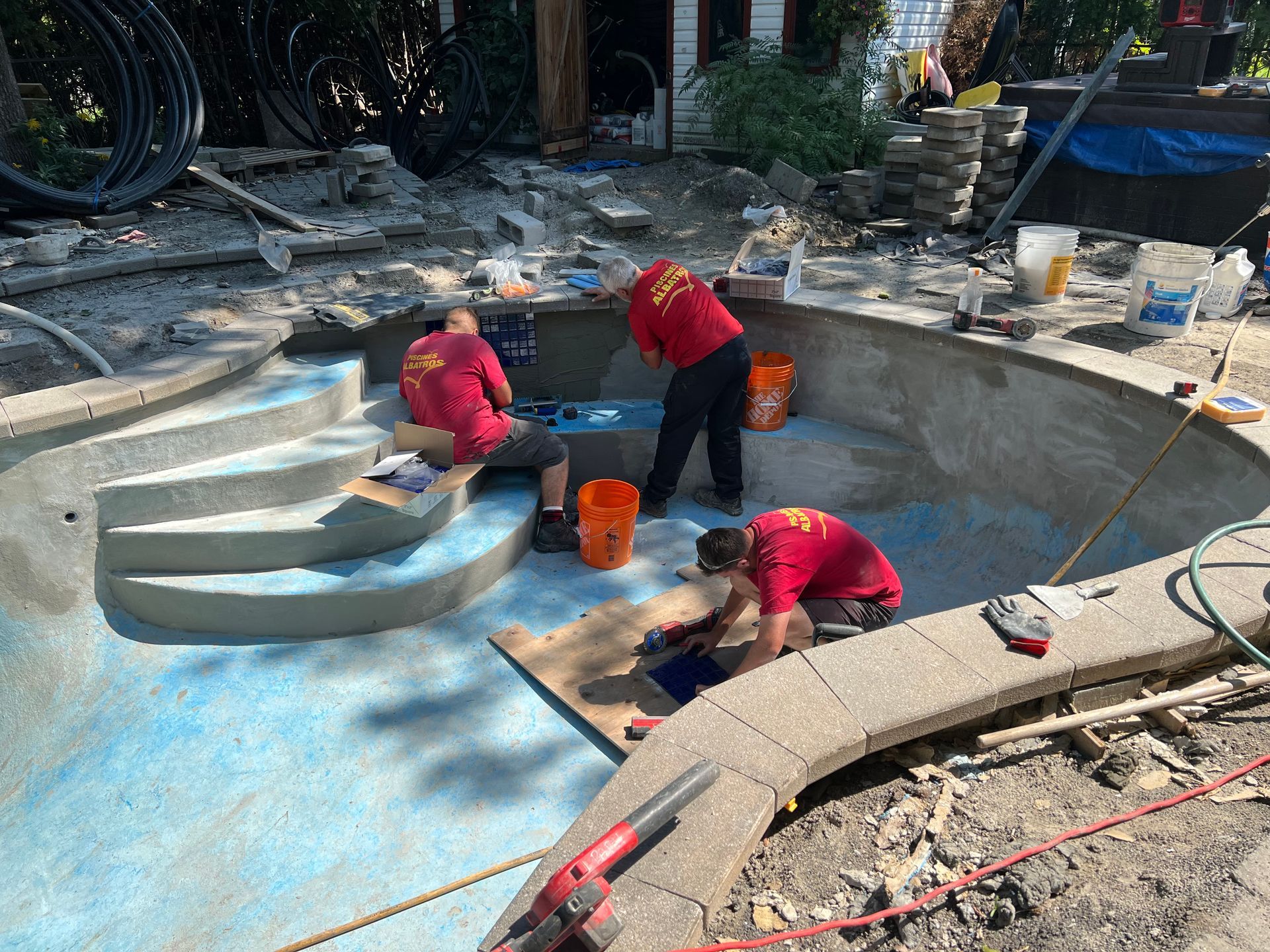 Trois ouvriers en chemises rouges travaillent à la finition des marches et du sol en béton à l'intérieur d'une piscine vide, peinte en bleu, située dans une cour arrière.