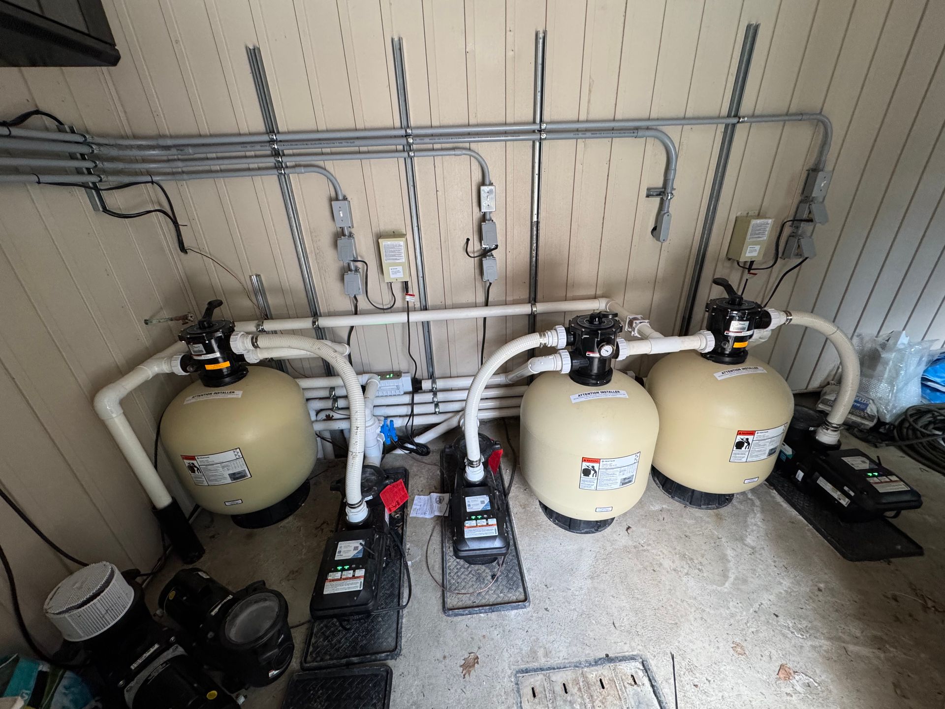 Trois systèmes de filtration de piscine avec pompes et réservoirs beiges sont disposés contre un mur comportant des conduits et des prises électriques.