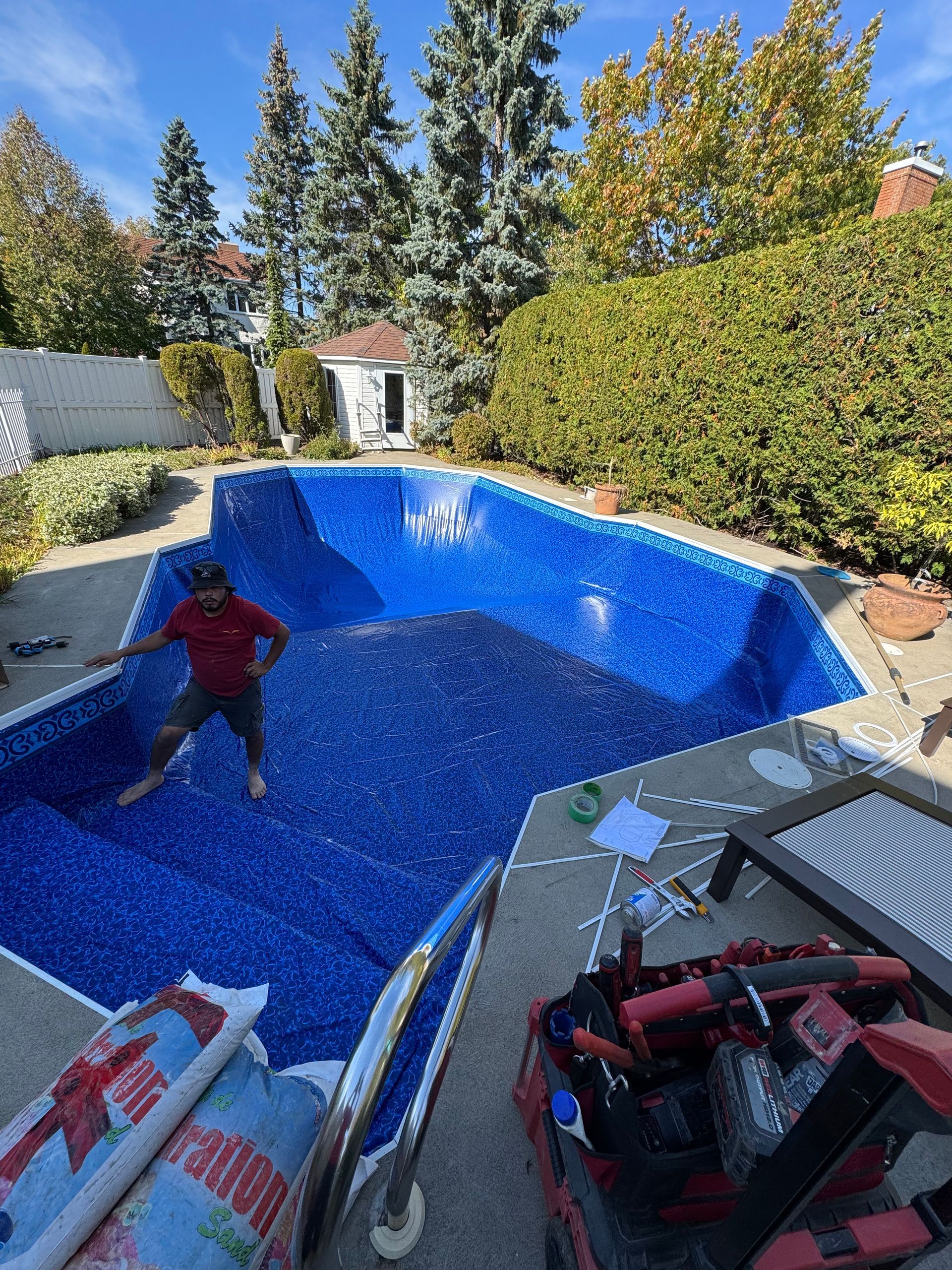 Piscine en rénovation dans le jardin, recouverte d'une bâche bleue ; un ouvrier se tient dans l'eau et des outils sont à proximité.