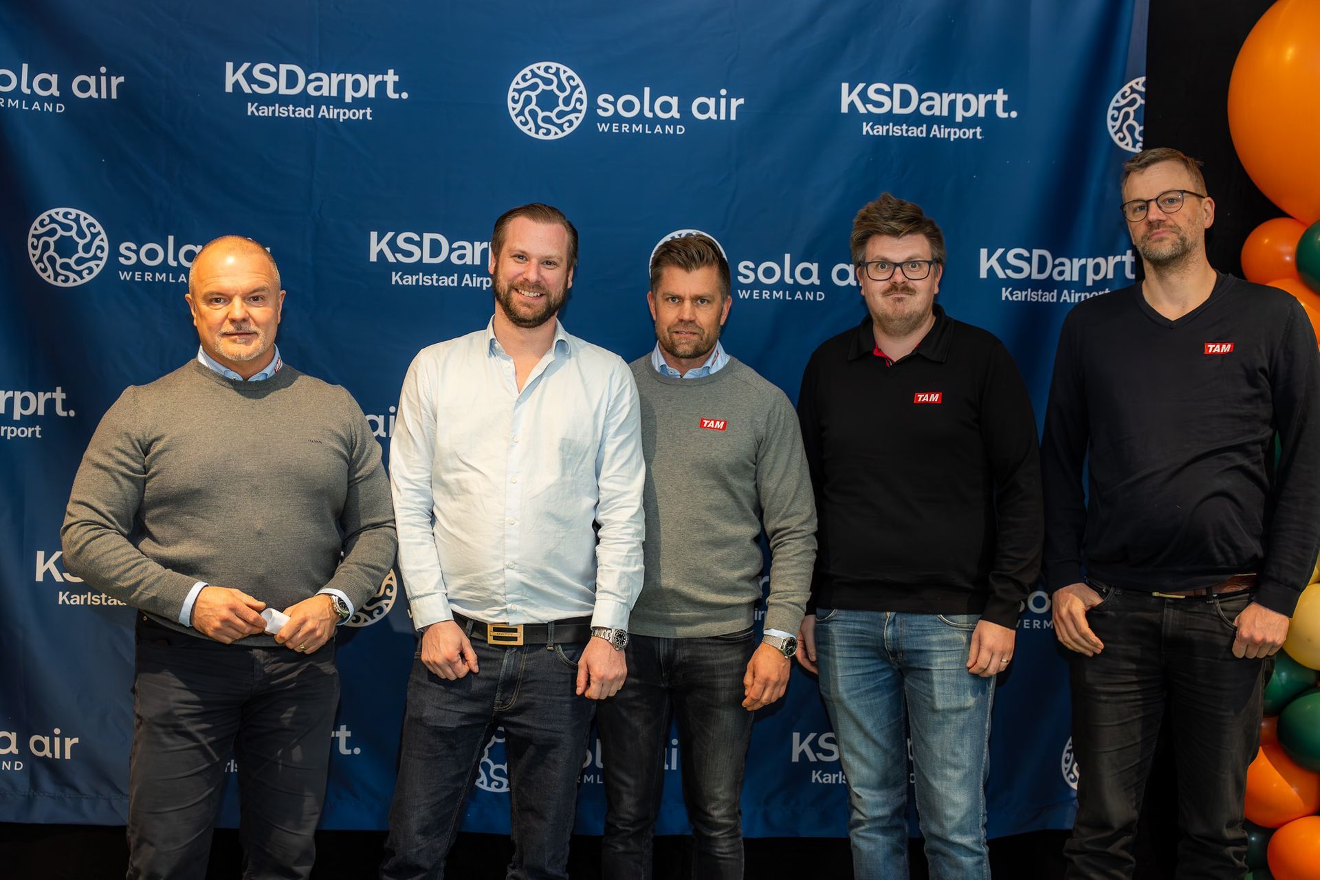 Fem män står vid en märkesdesignad step-and-repeat-evenemangsbakgrund och poserar för ett gruppfoto