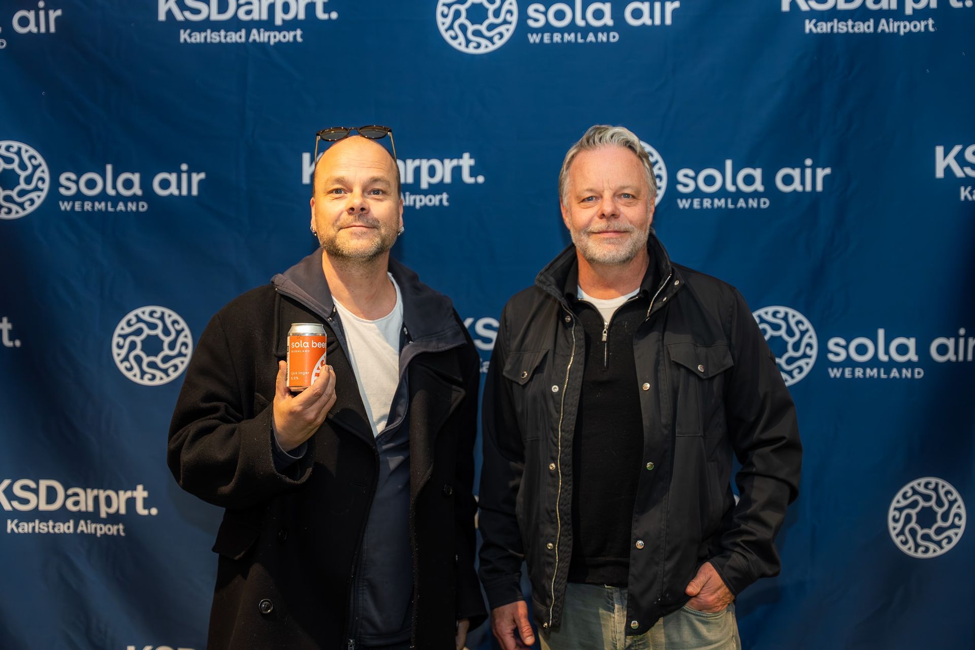 Två män poserar mot en Sola Air-bakgrund, en håller en burk, båda ler nonchalant