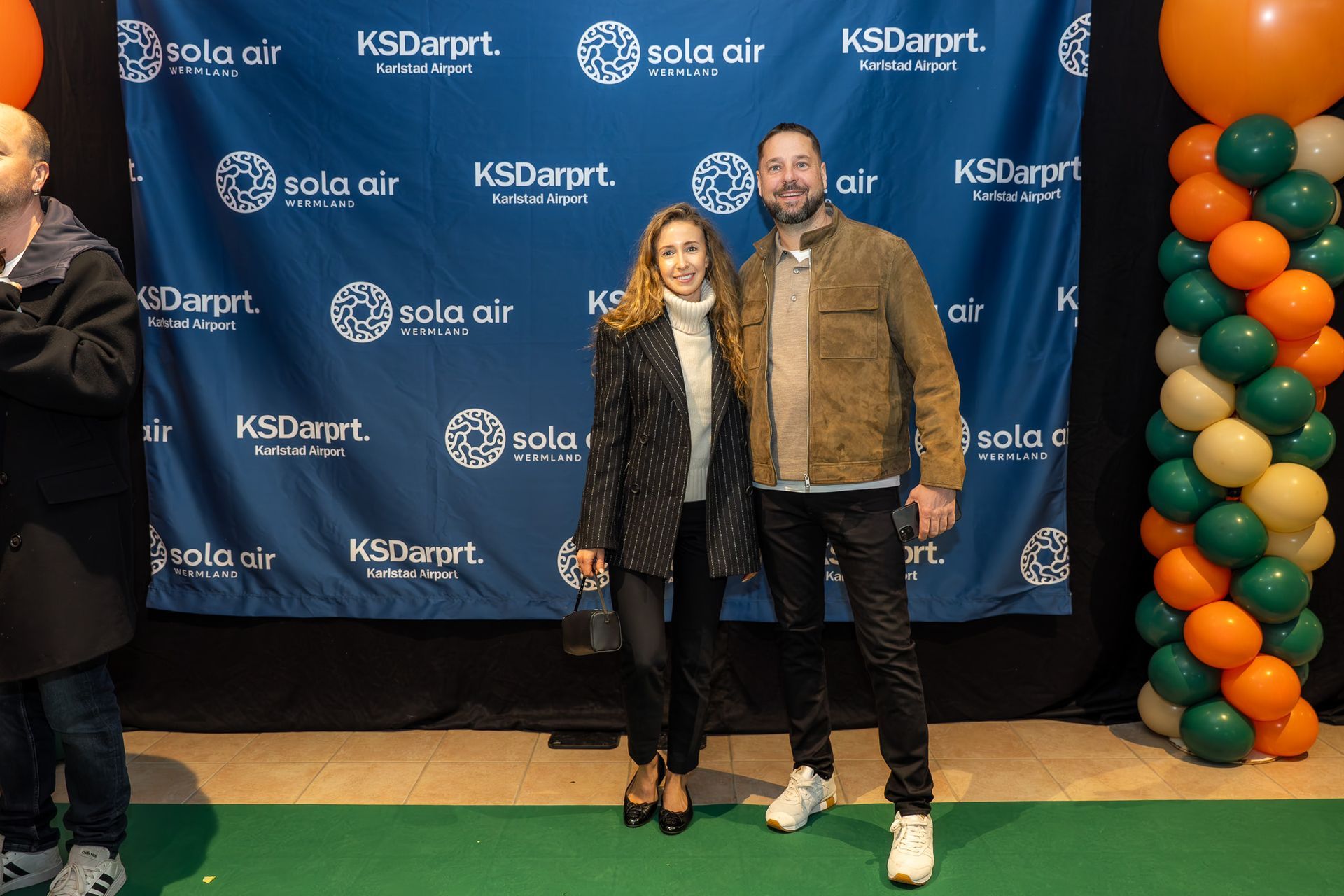 Två personer poserar framför en KSDart/Sola Air-bakgrund med orange och gröna ballongpelare