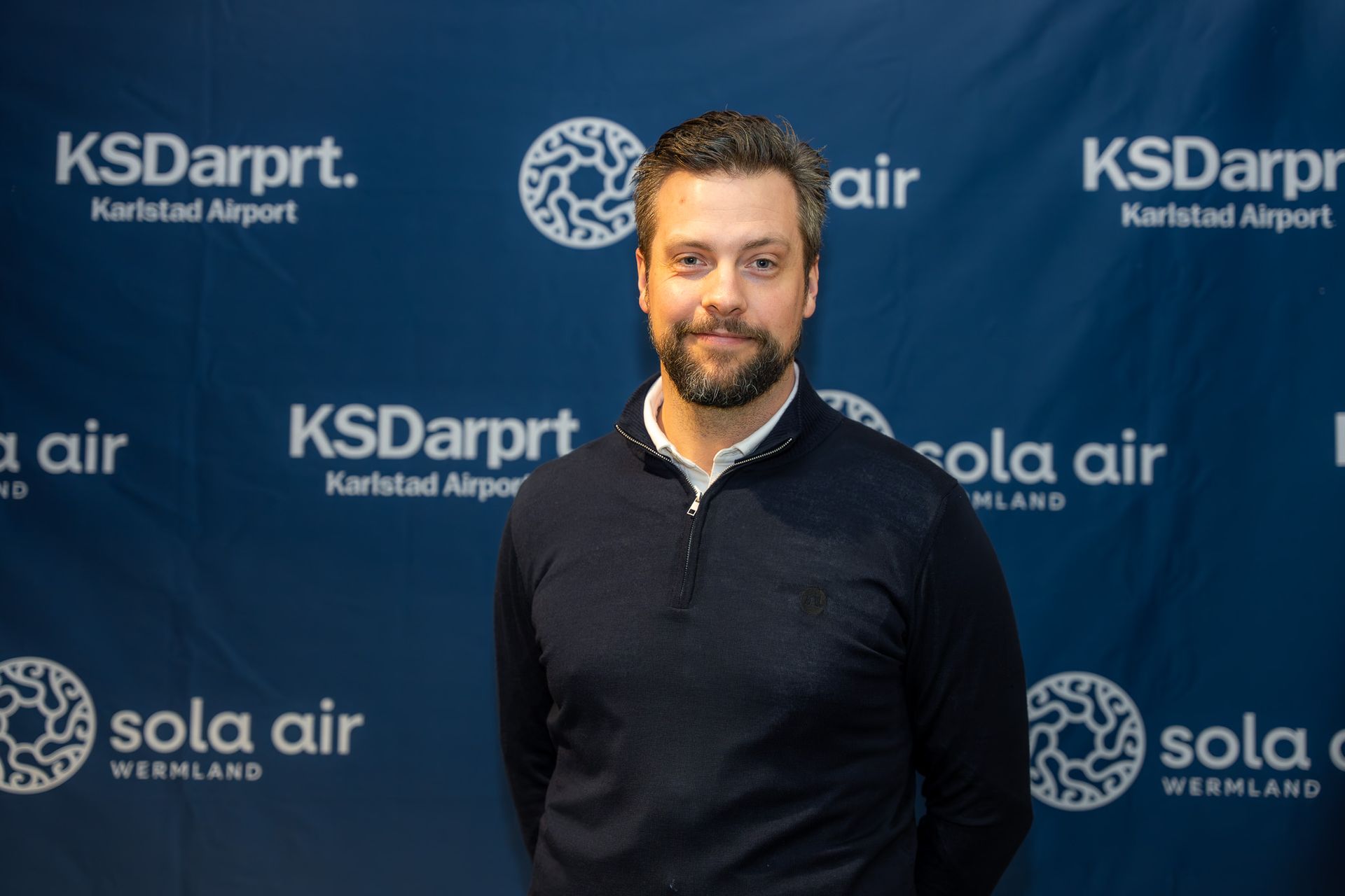 Man står framför en blå bakgrund med KS Darrpt- och Sola Air-märken, klädd i en mörk tröja och ler