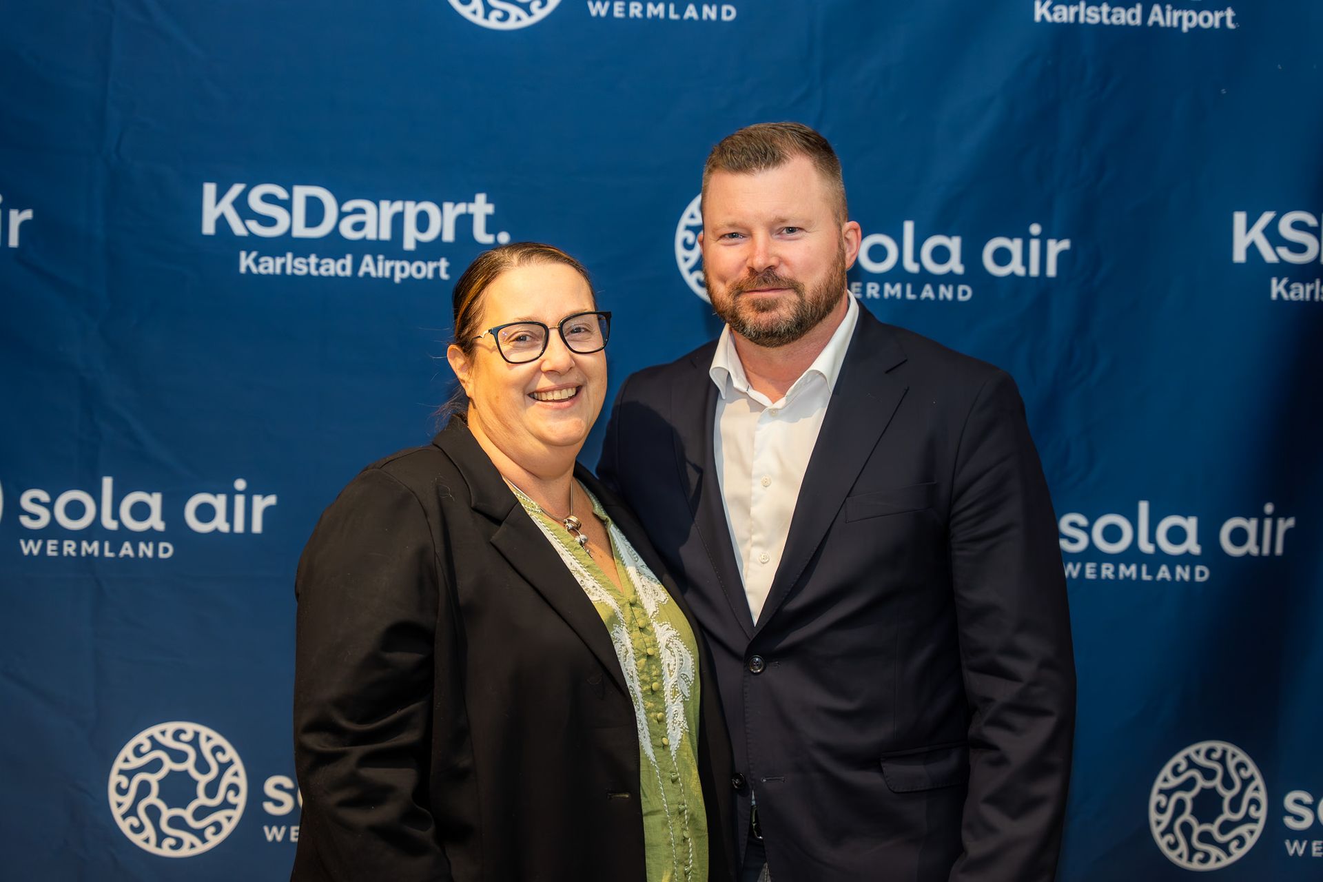 Två personer poserar leende framför en bakgrund med varumärket KSD Airport och Solair på ett evenemang