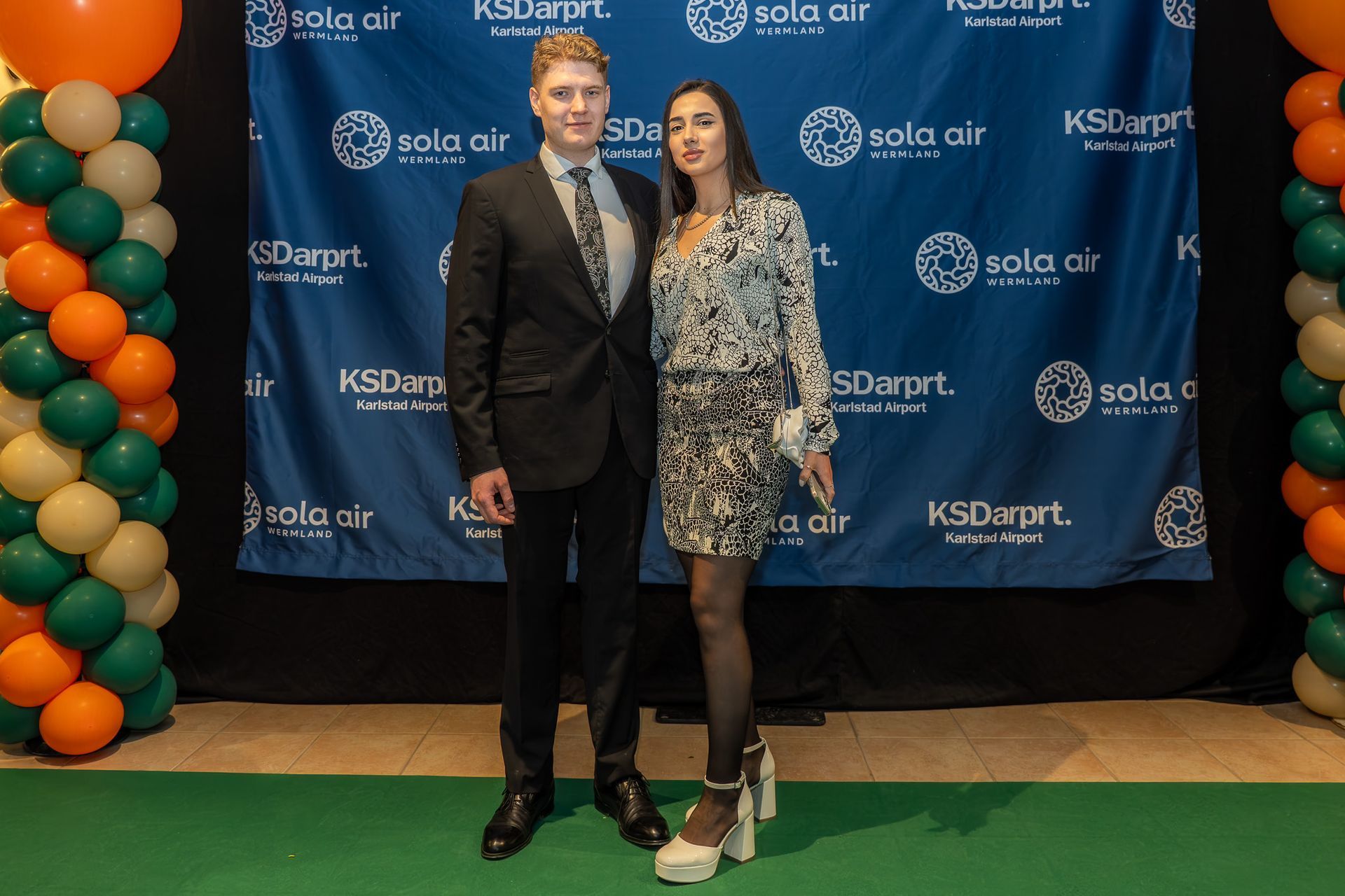 Två personer poserar på en grön matta framför en blå sponsorbakgrund och ballongbåge