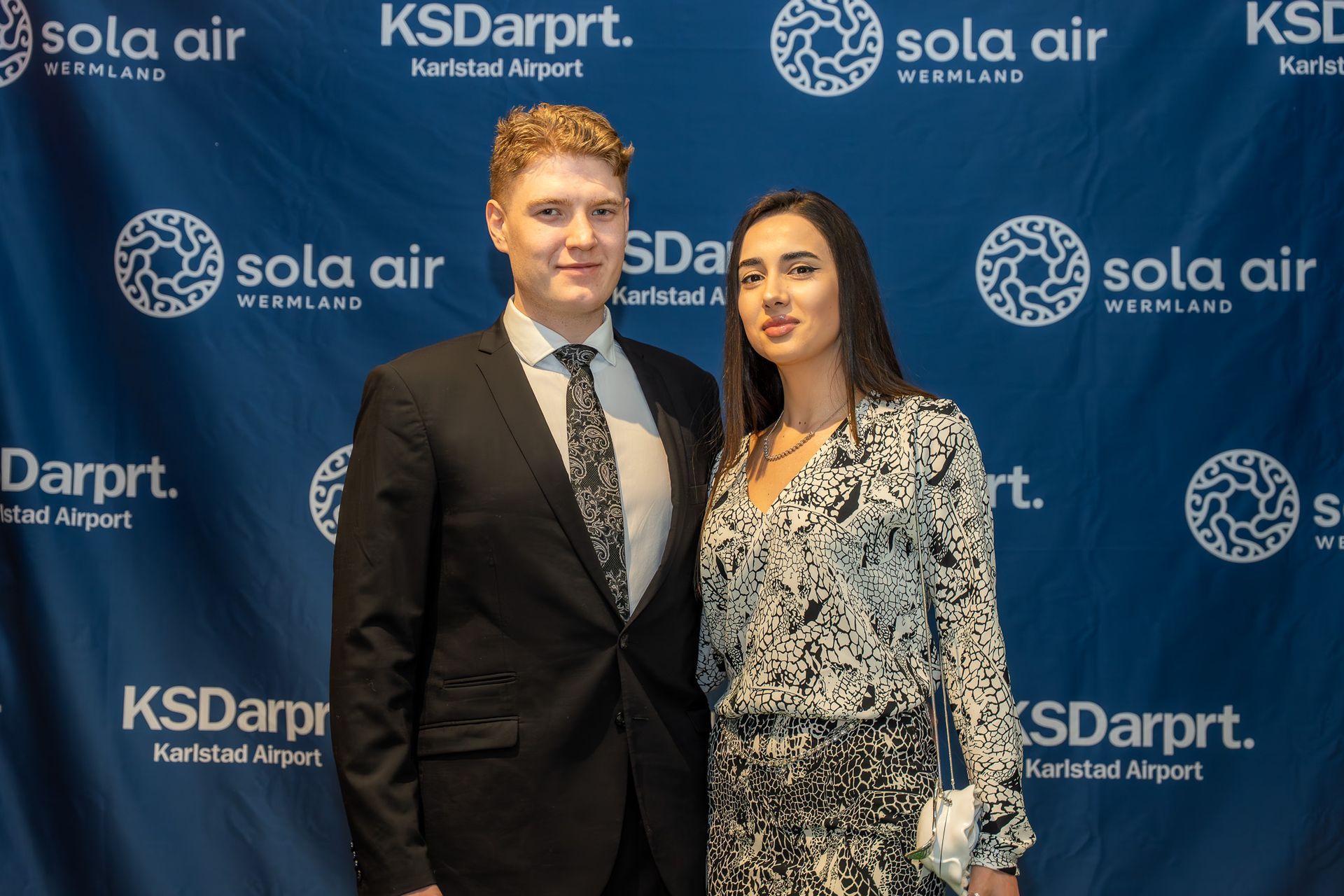Två personer poserar framför en blå Solo Air och KSDarpt. Karabat flygplats-bakgrund