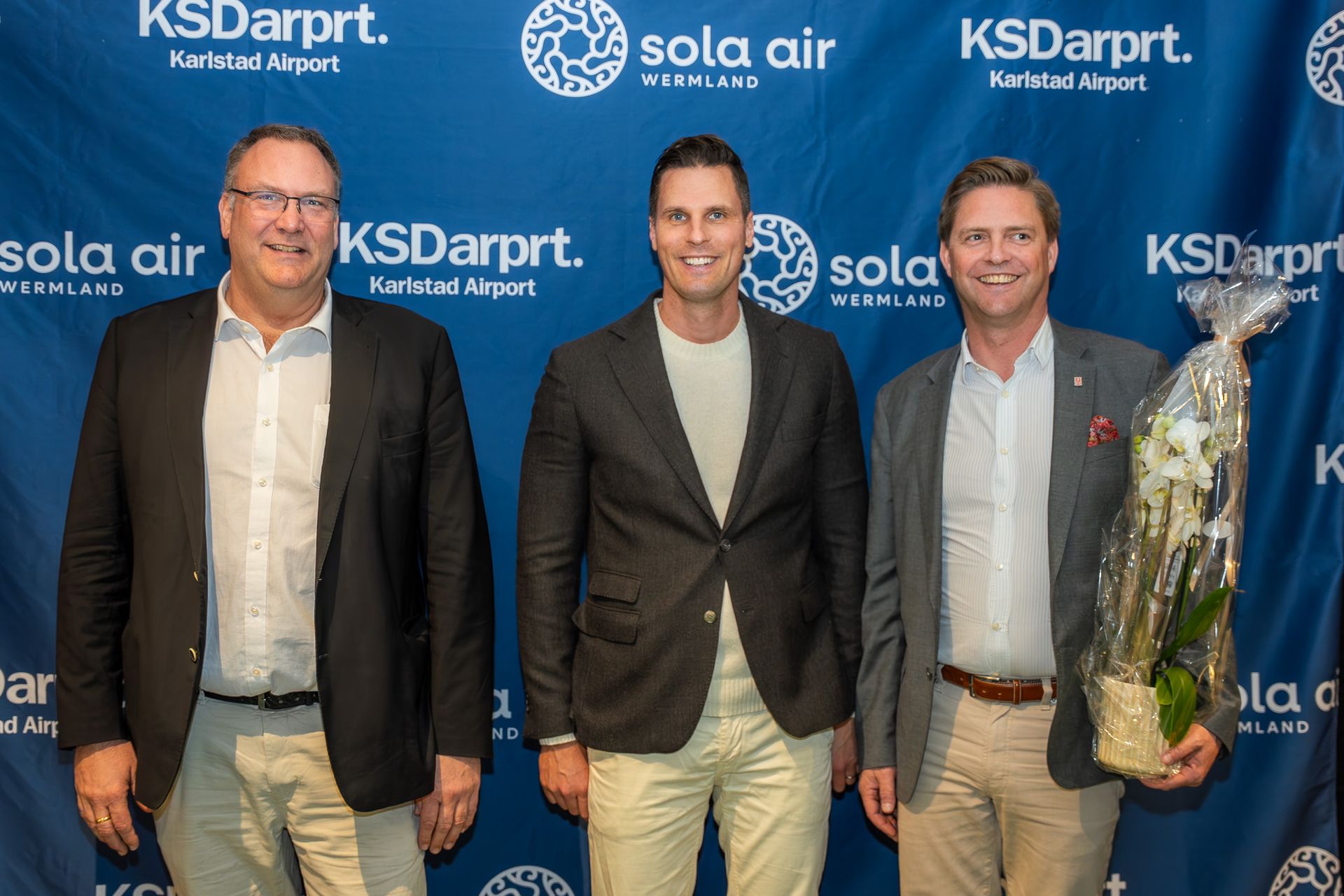 Tre män poserar vid en bakgrund av ett KSDarprt- och Sola Air-evenemang, med en inslagen presentkorg i handen.