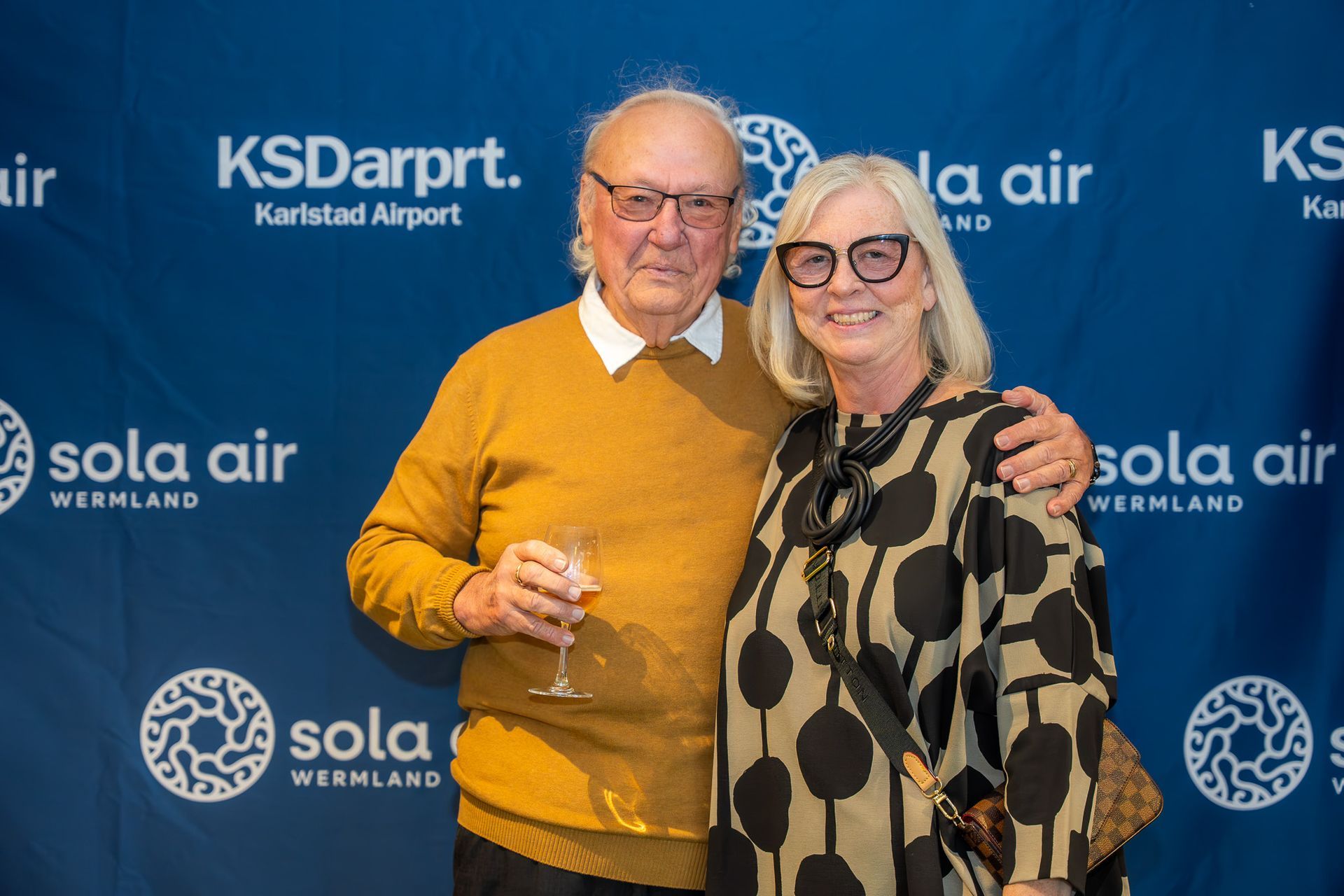 Två leende personer poserar med drinkar i bakgrunden av ett KSDart- och Sola-flygevenemang