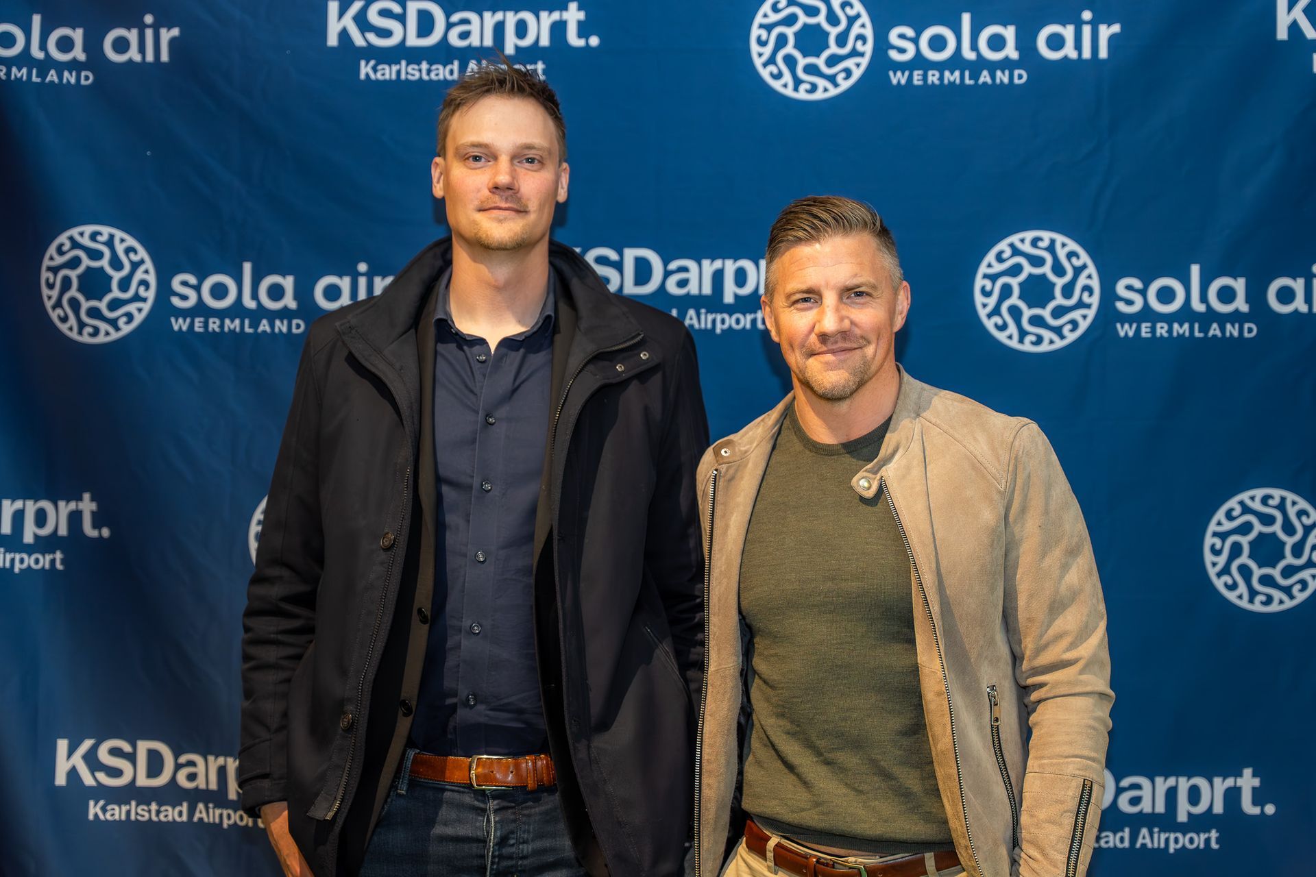 Två män poserar framför en blå KSDarprt. och Sola Air-bakgrund vid ett evenemang.