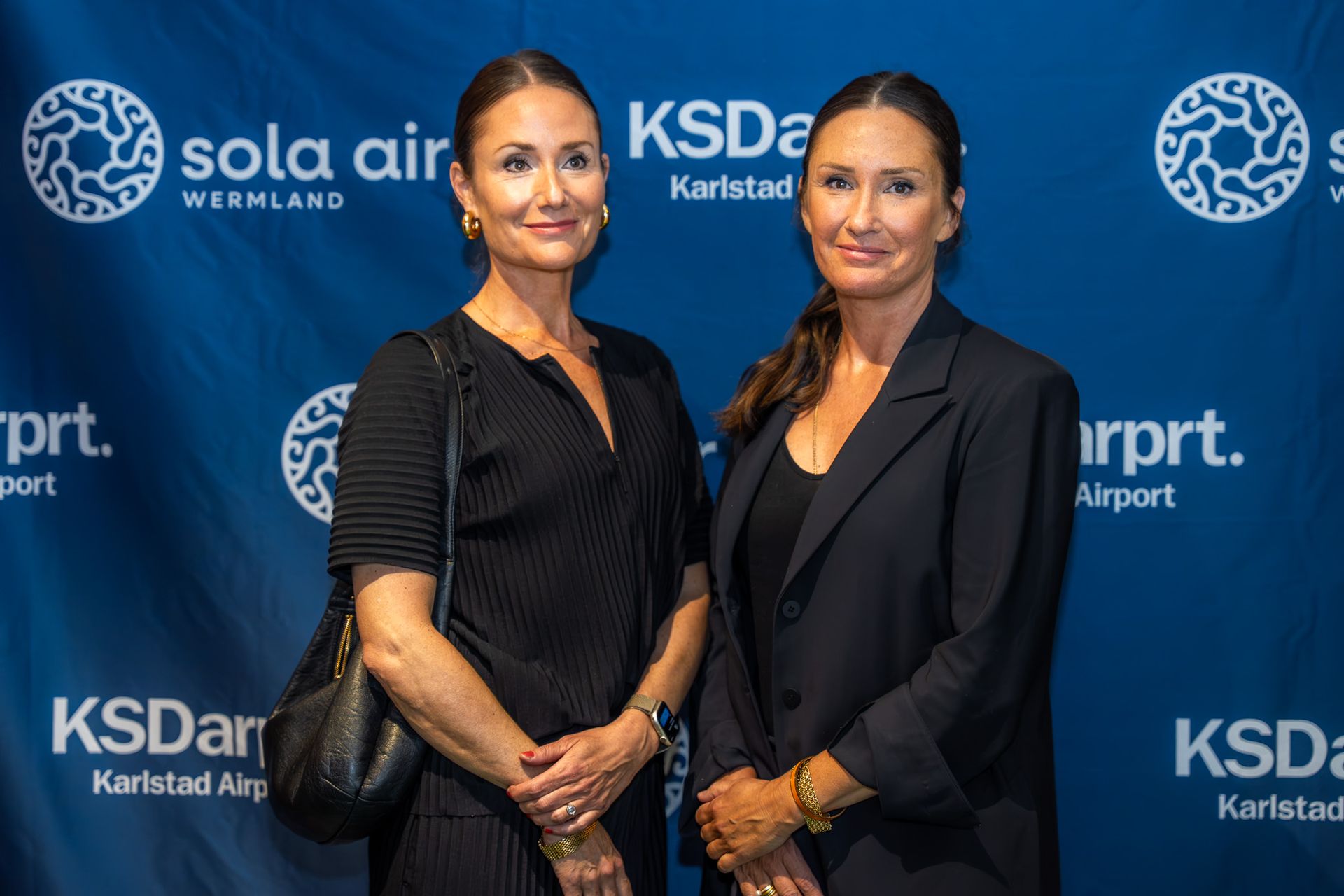 Två kvinnor i svarta kläder poserar inför en blå evenemangsbakgrund med logotyper