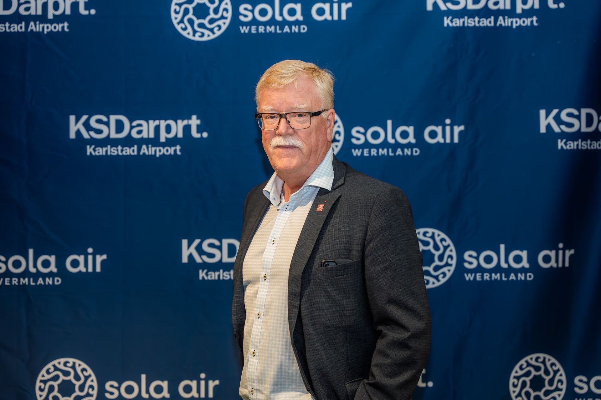 Man står framför en blå Sola Air- och KSDarpt-bakgrund, iklädd kavaj och glasögon.