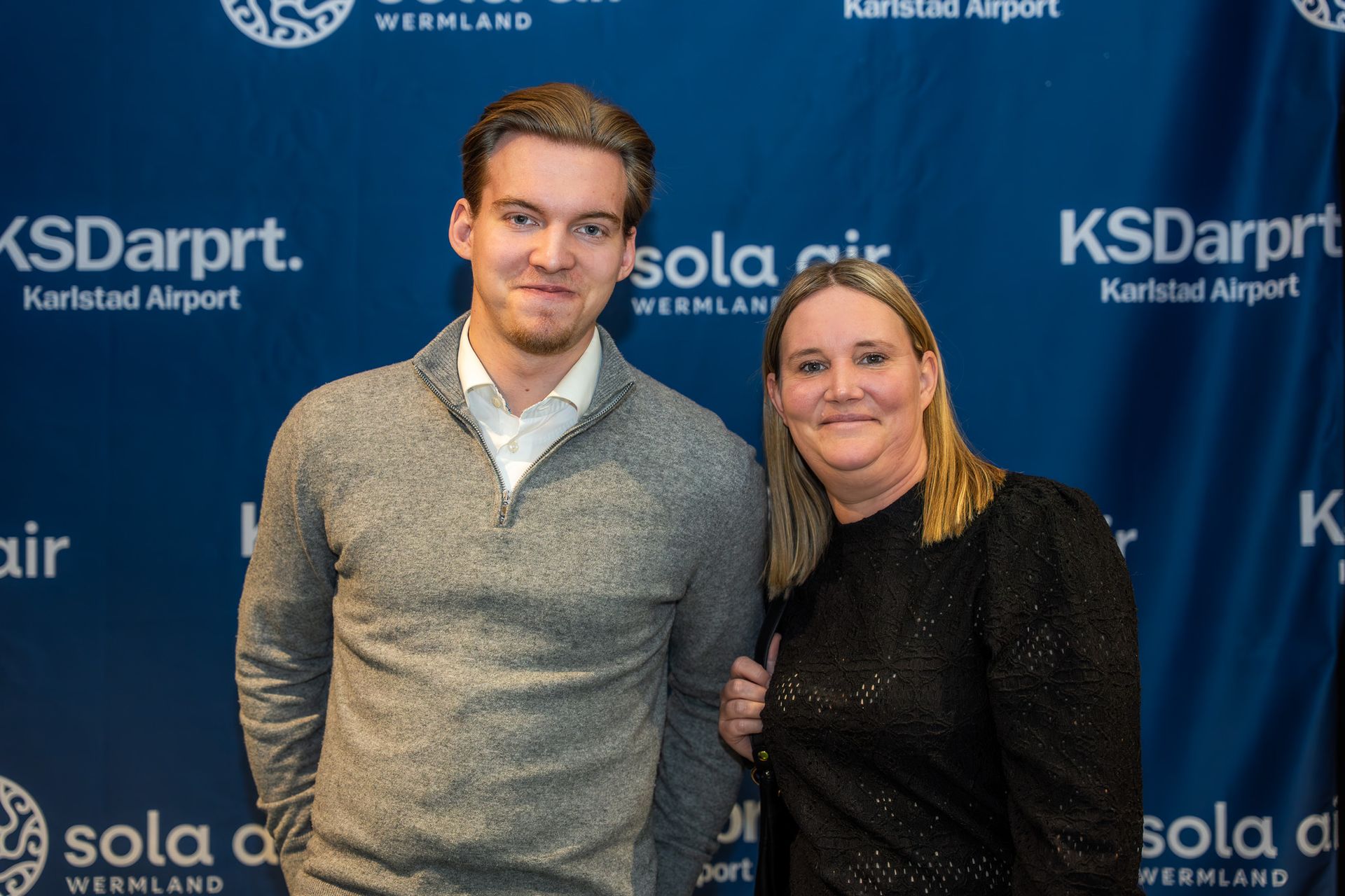 Två leende personer poserar framför en blå evenemangsbakgrund med logotyperna ”KSDarpt. Karlstad Airport”.