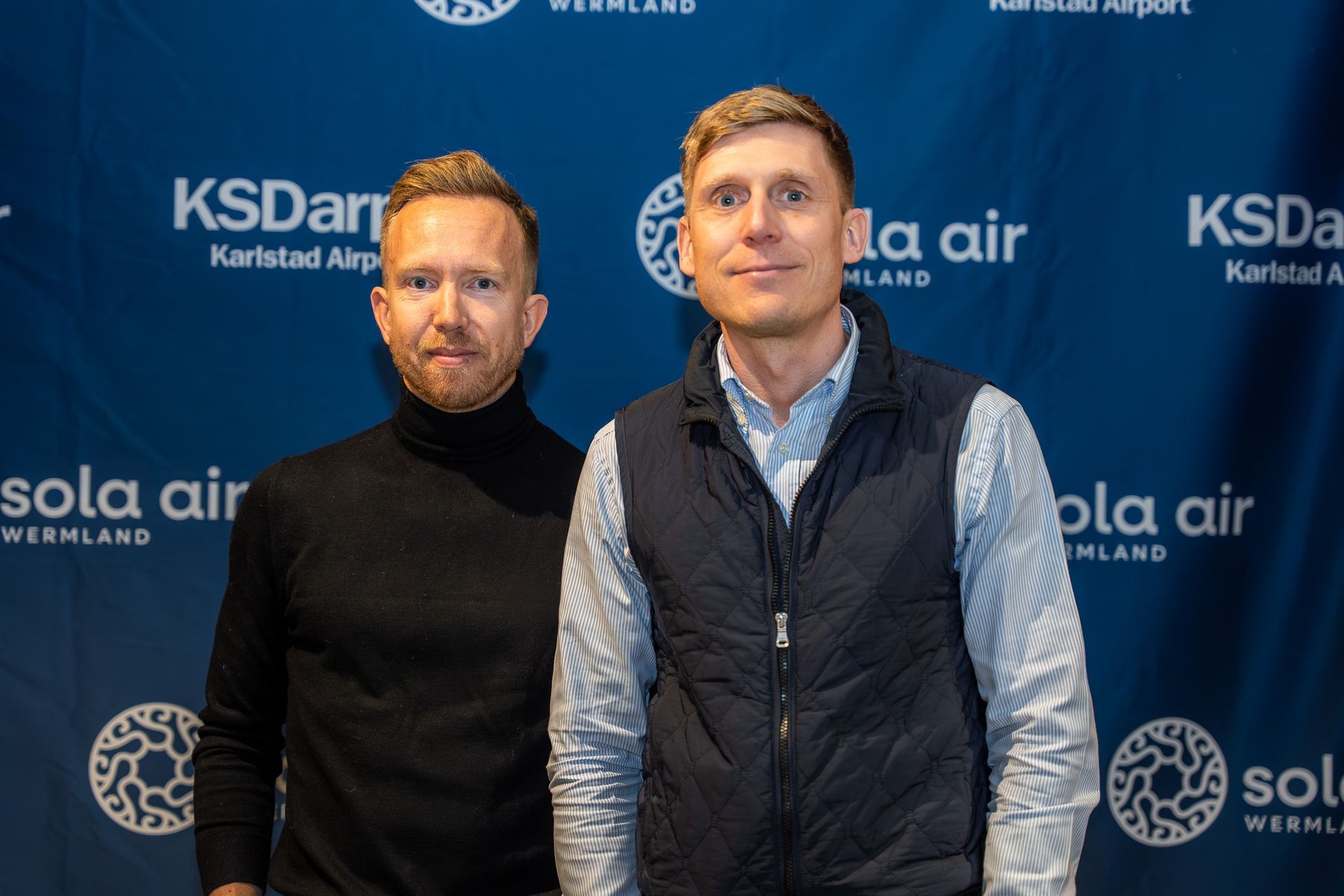 Två män står framför en blå KSDartsola Air-kuliss vid ett evenemang, en i svart polotröja och en i en marinblå väst