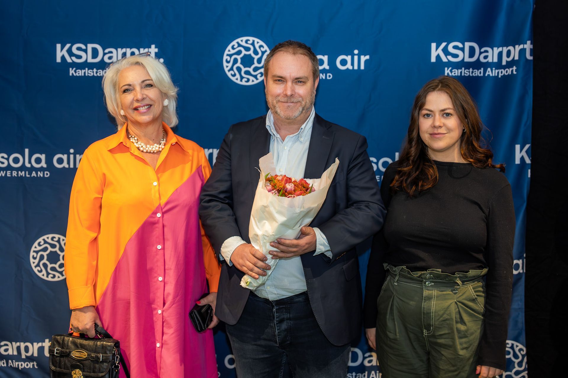 Tre personer poserar på ett KSDarprrt och Sola Air-evenemang, varav en håller en blombukett.