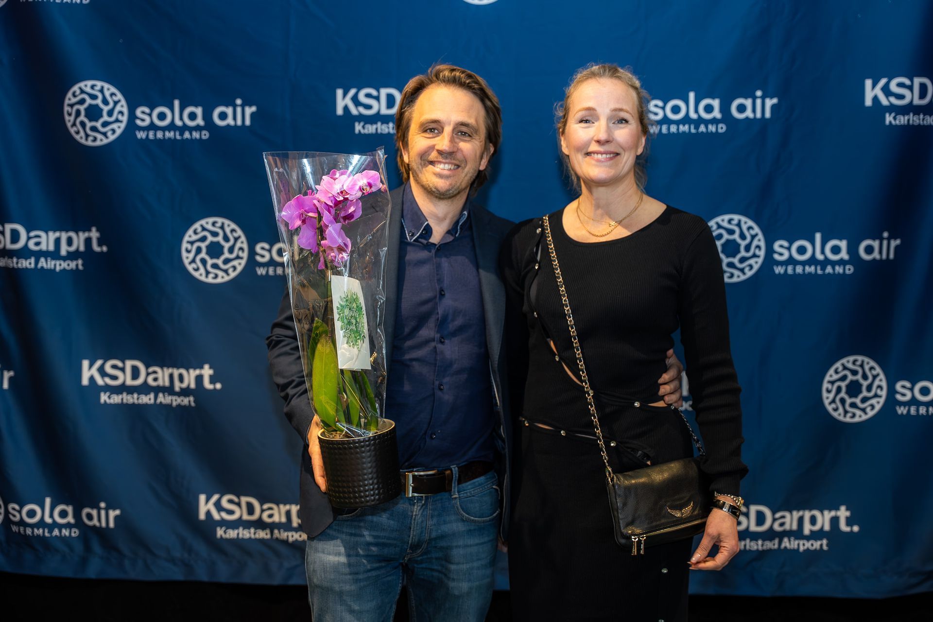 Två personer poserar leende vid en bakgrund av ett KSDartp-evenemang, en av dem håller rosa blommor.