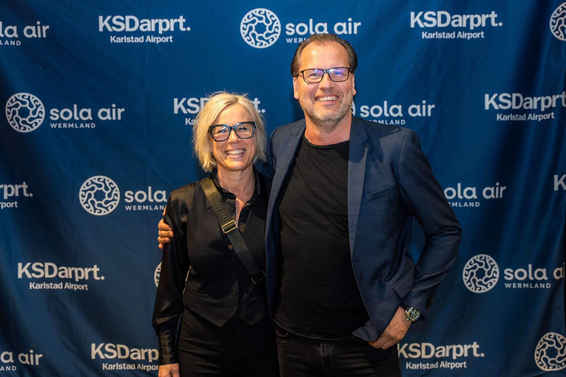 Två personer poserar leende framför en blå bakgrund med logotyper för ett step-and-repeat-evenemang.