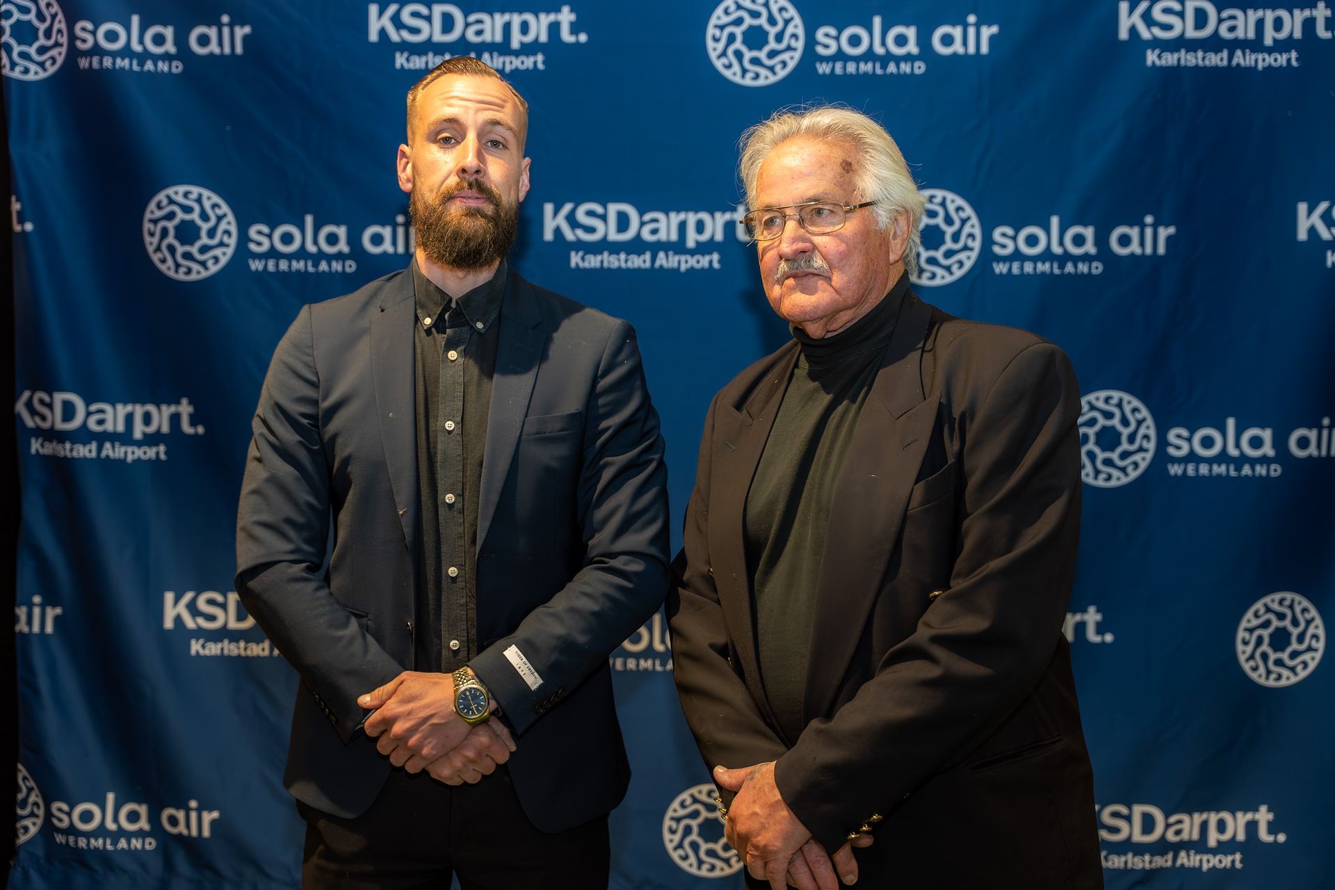 Två män poserar framför en blå bakgrund med KSDarput- och Sola Air-logotyper vid ett formellt evenemang