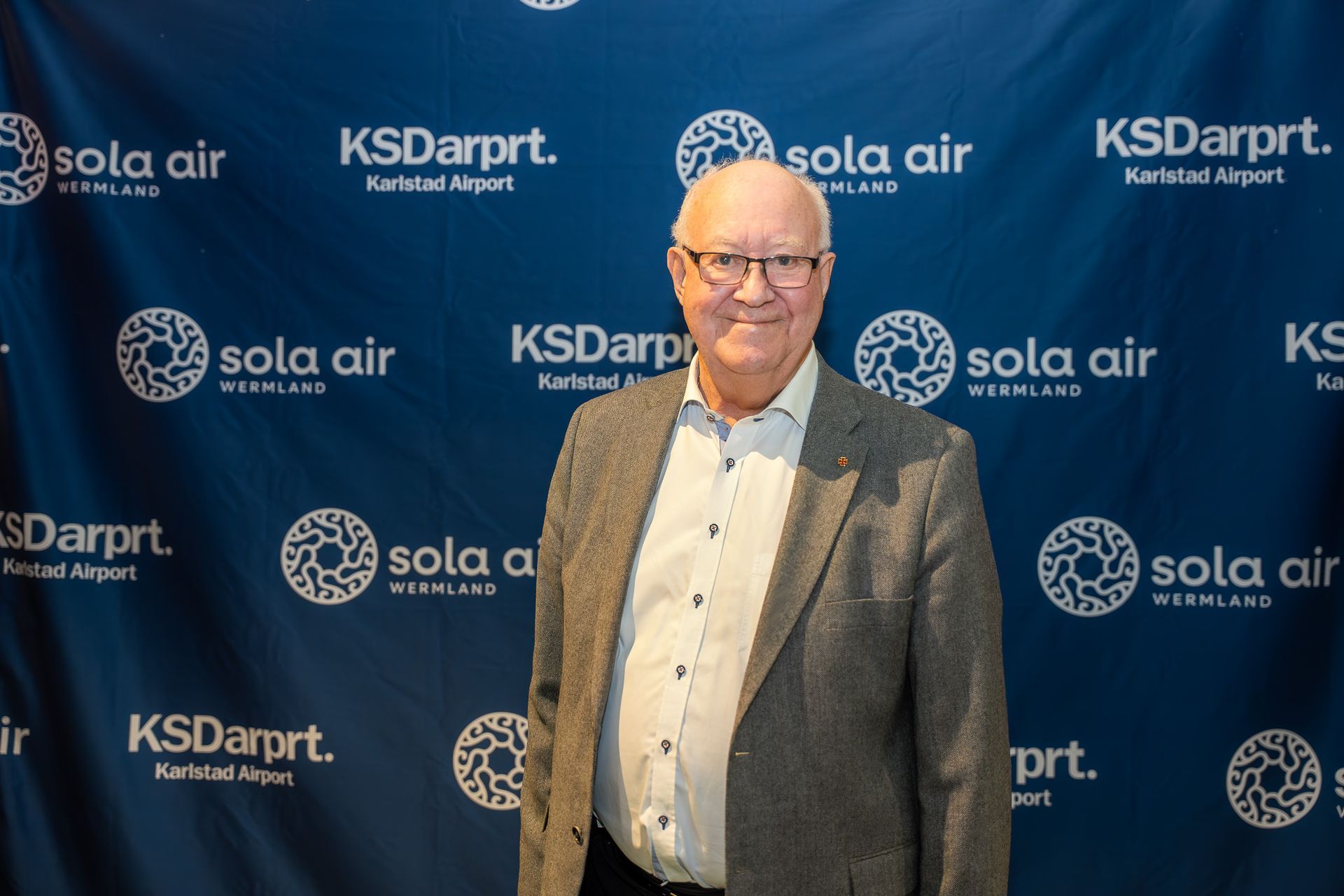 Man i kavaj och fluga poserar vid en evenemangsbakgrund med logotyperna ”Sola Air” och ”KSDarput”