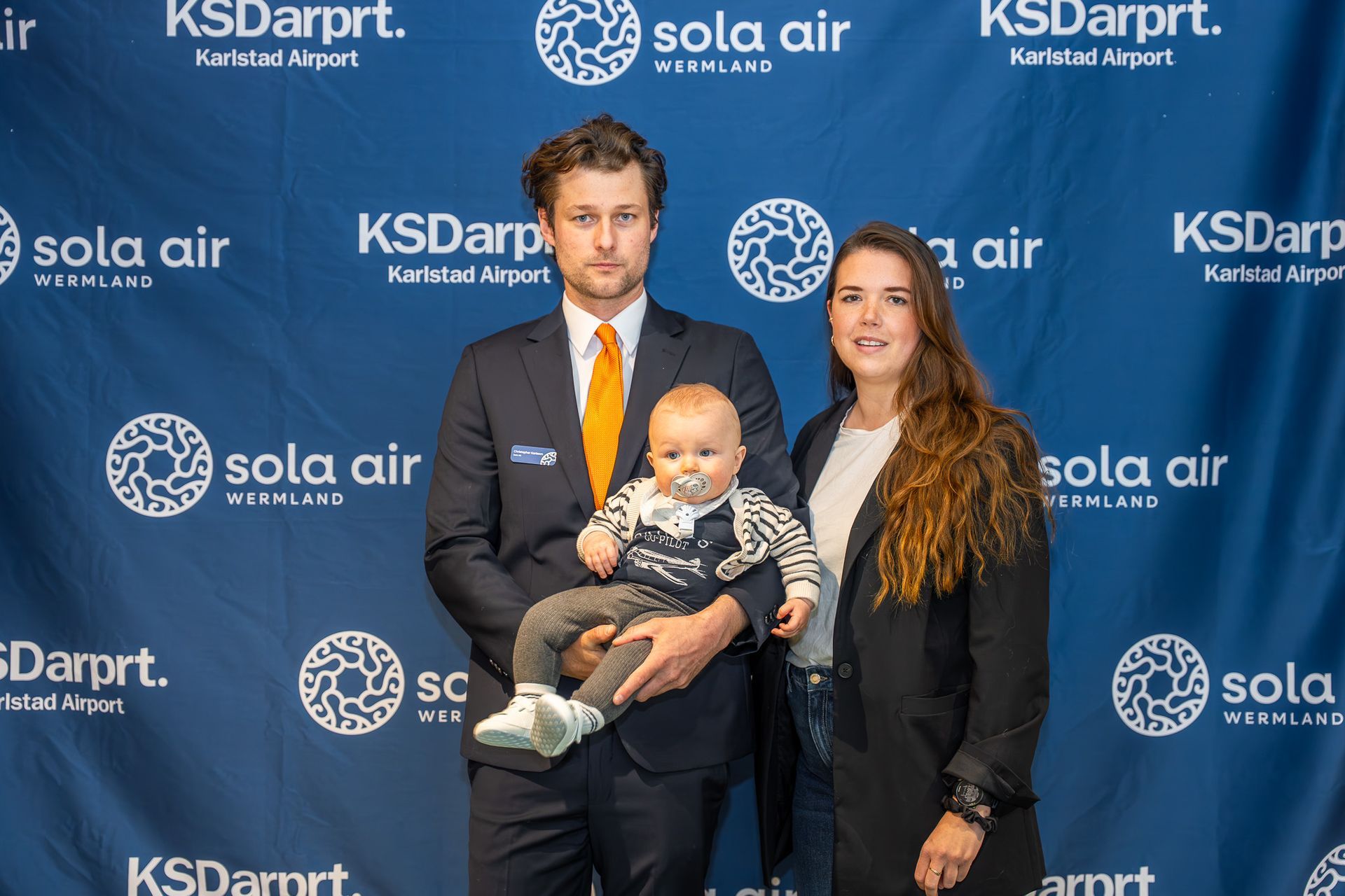 Tre personer poserar i bakgrunden av ett KSDarpt- och Sola Air-evenemang med en bebis i handen.