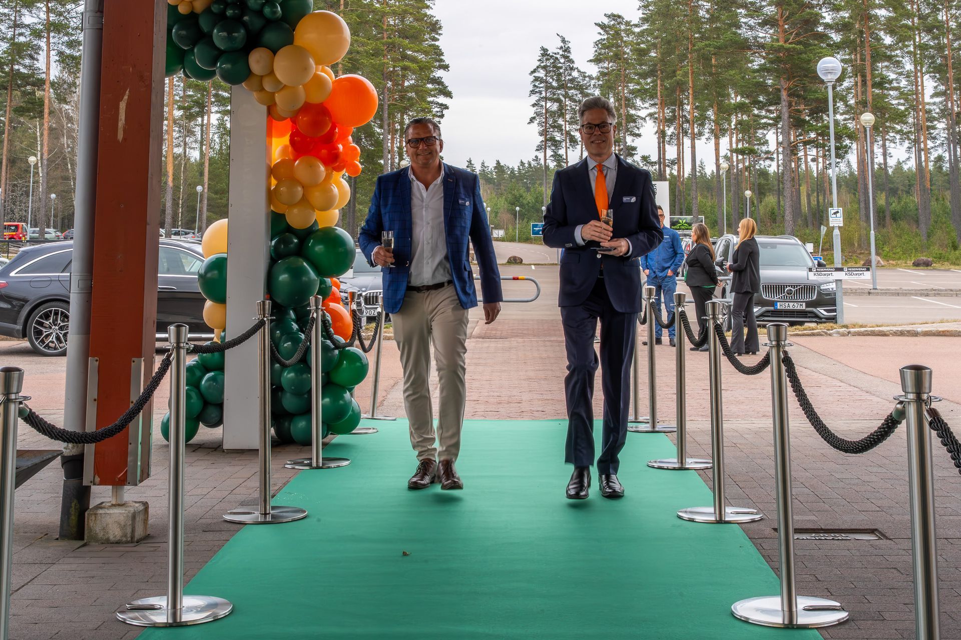 Två män som går på en grön mattbelagd entré med ballonger och ett repbarriär, utomhus nära träd