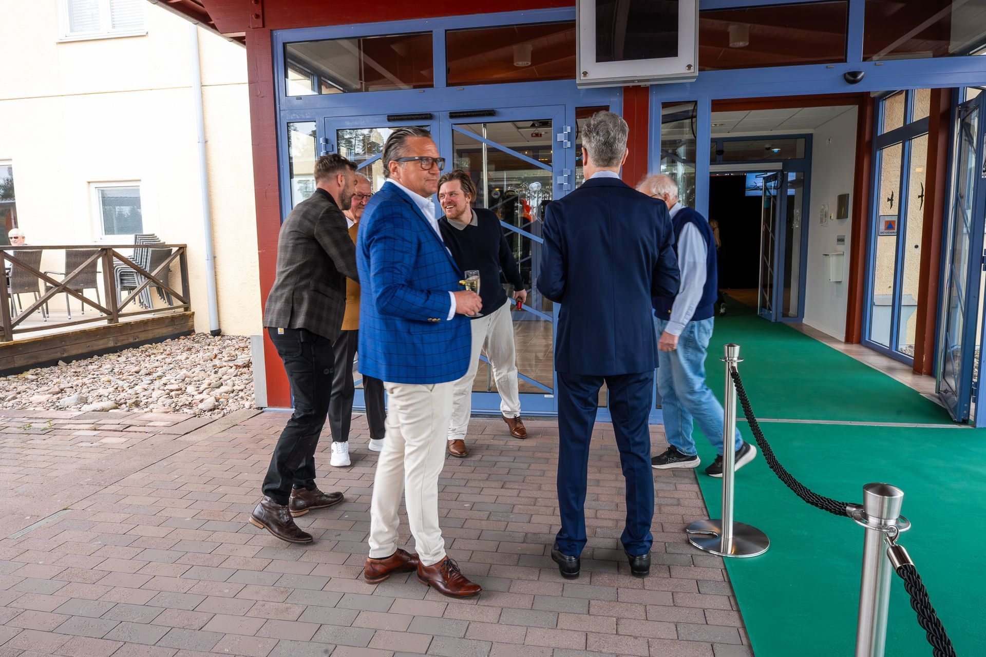 Grupp människor som står utanför en byggnads entré på en grön matta och pratar nära dörröppningen.