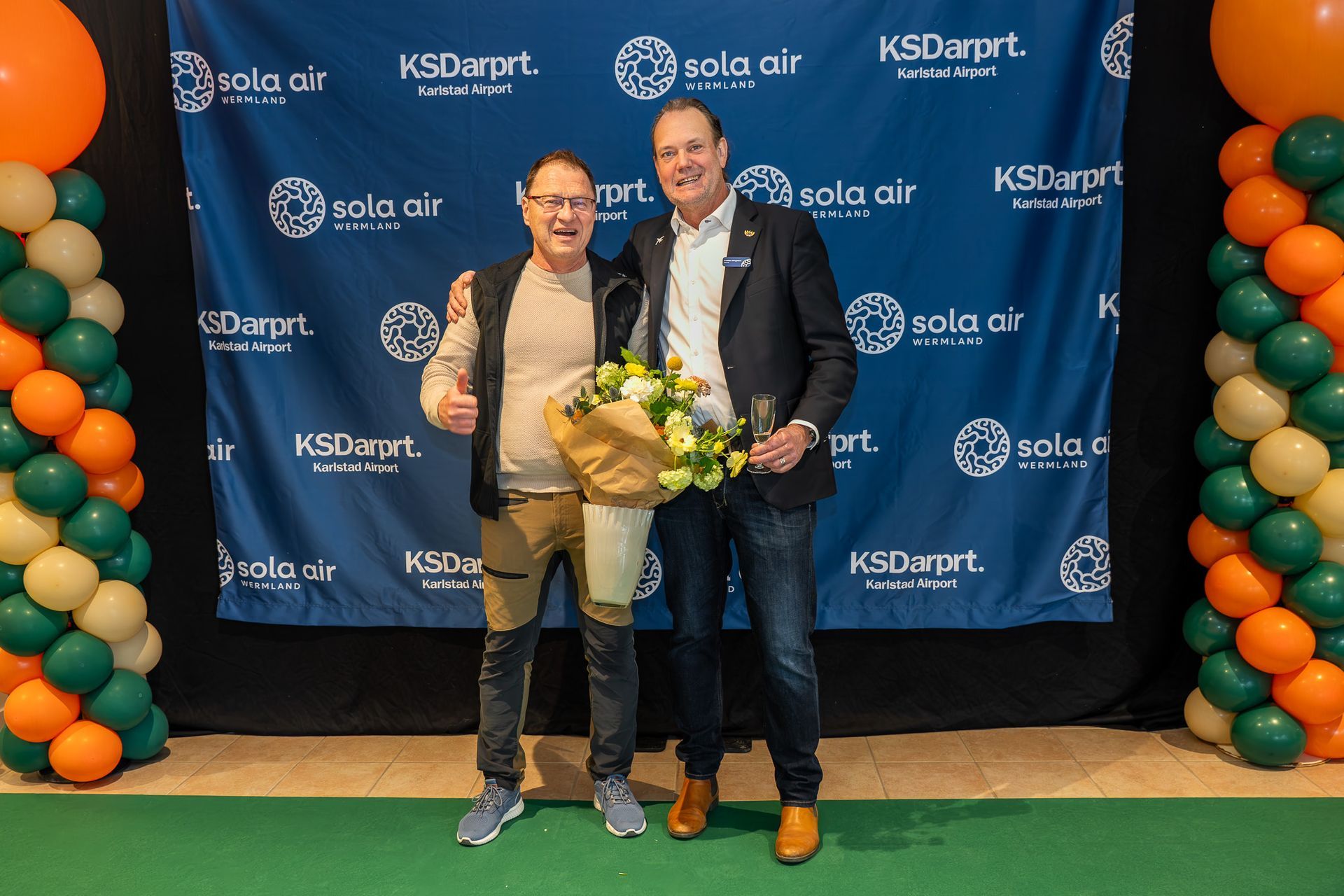 Två personer som håller blommor poserar framför en KSD Events steg-och-repeat-bakgrund med ballongpelare.