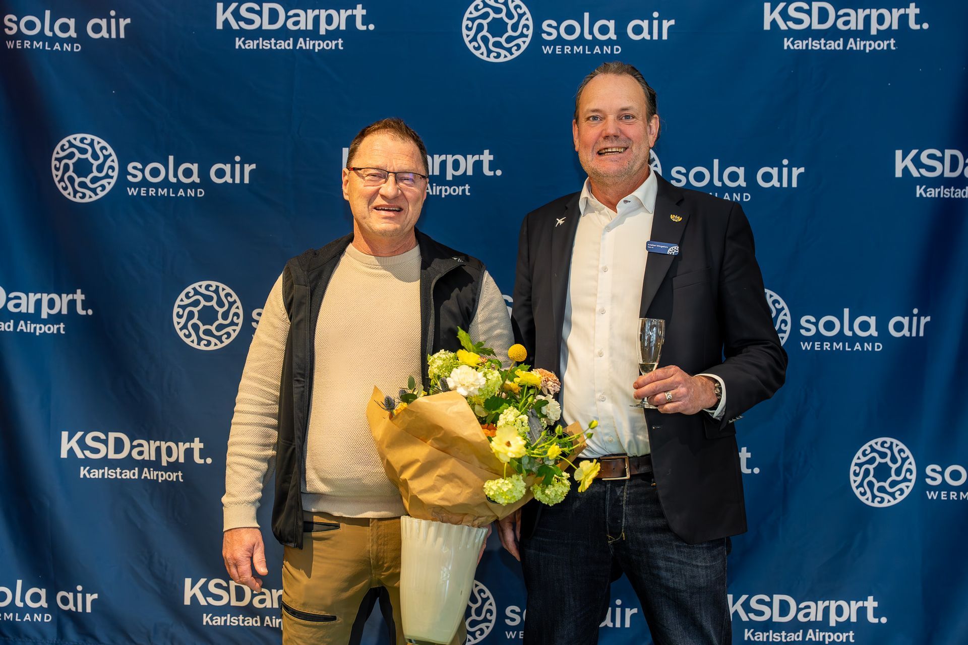 Två män som ler med en bukett mot en Sola Air och KSDarpt-bakgrund.