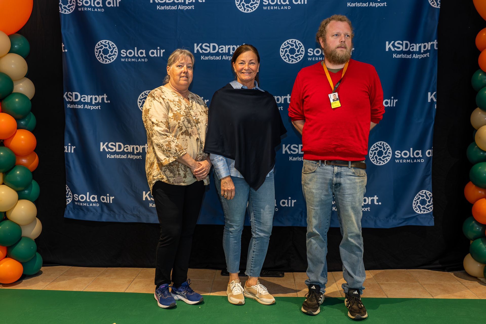 Tre personer står framför en märkesbakgrund på ett evenemang och poserar för ett foto