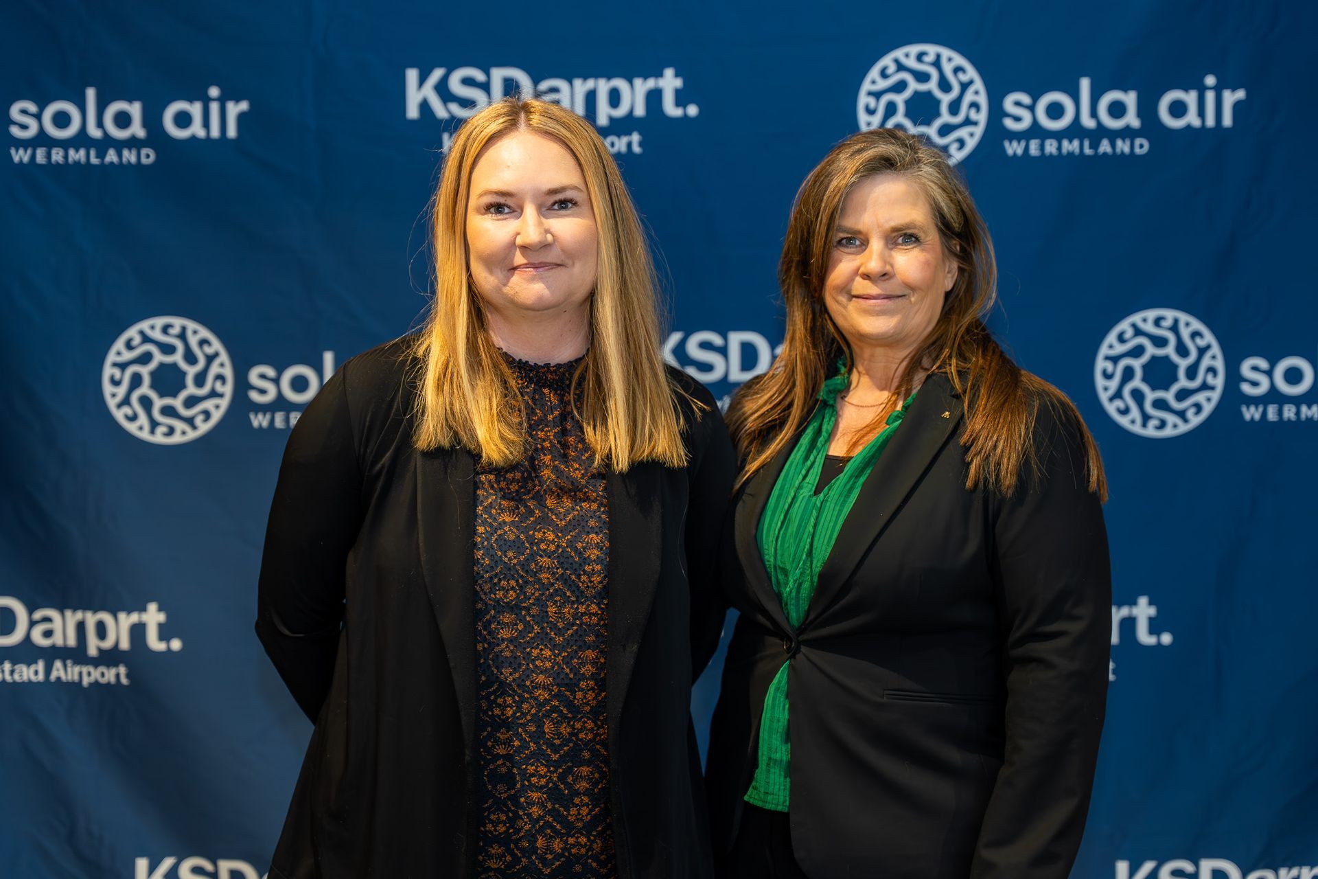 Två kvinnor poserar vid ett evenemang på KSD:s flygplats i bakgrunden med Sola Air- och KSDarpt-logotyper.