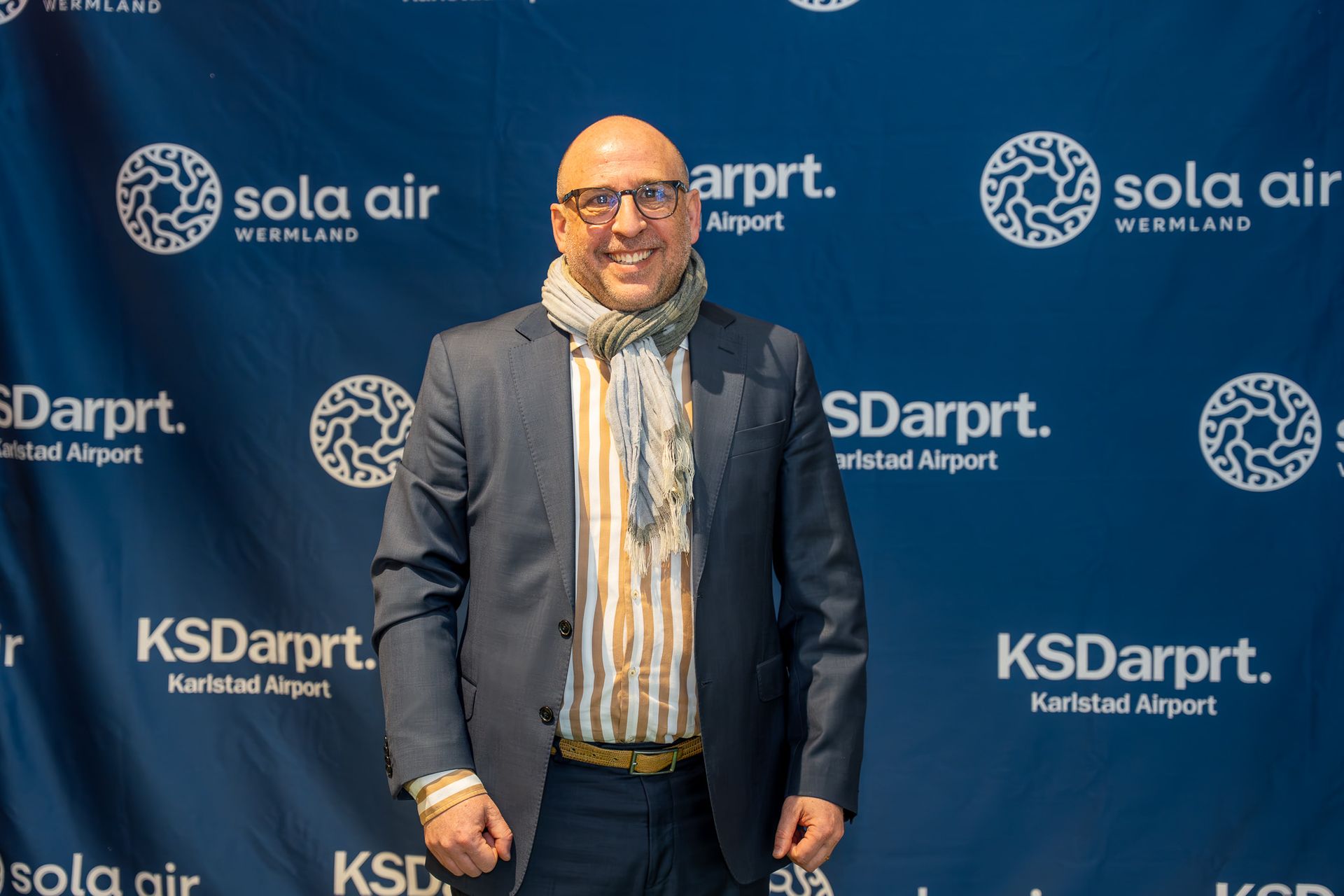 Man i mörk kavaj och halsduk poserar inför en blå bakgrund av en KSDarpt-flygplatssponsor