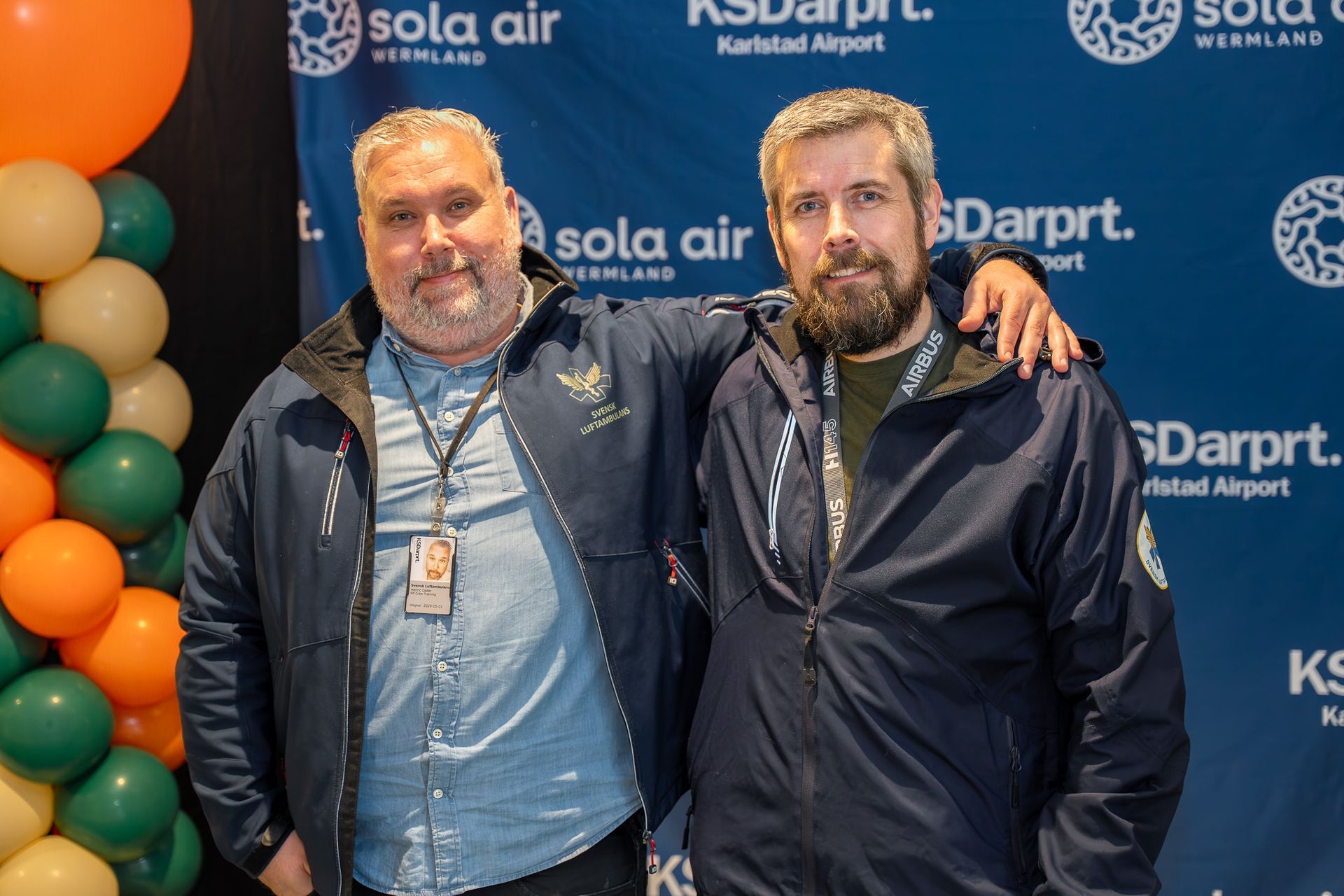 Två män poserar tillsammans mot en Sola Air-bakgrund, med ballonger till vänster.