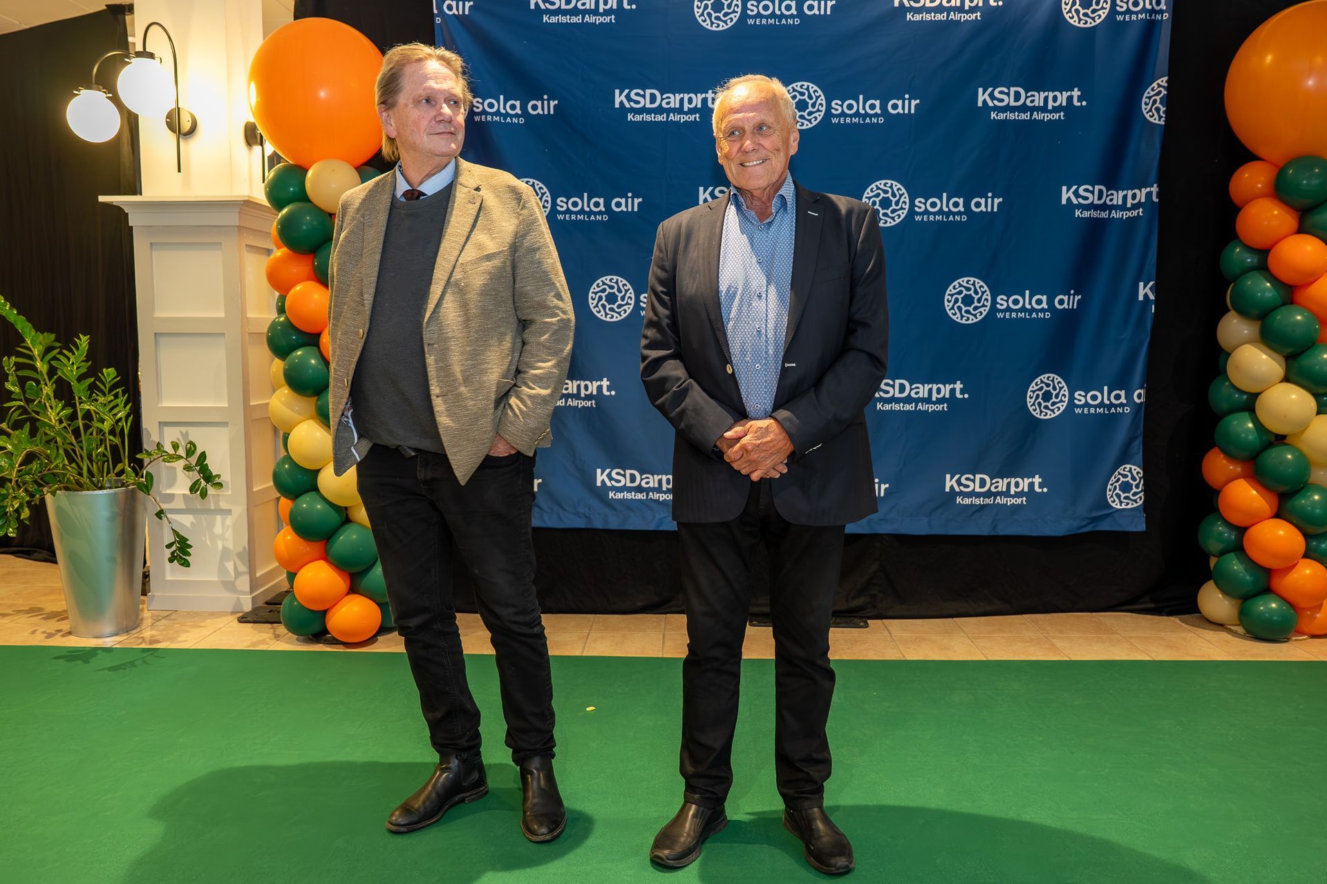 Två män i jackor står på en grön matta vid ett evenemang med orange och svarta ballongdekorationer.