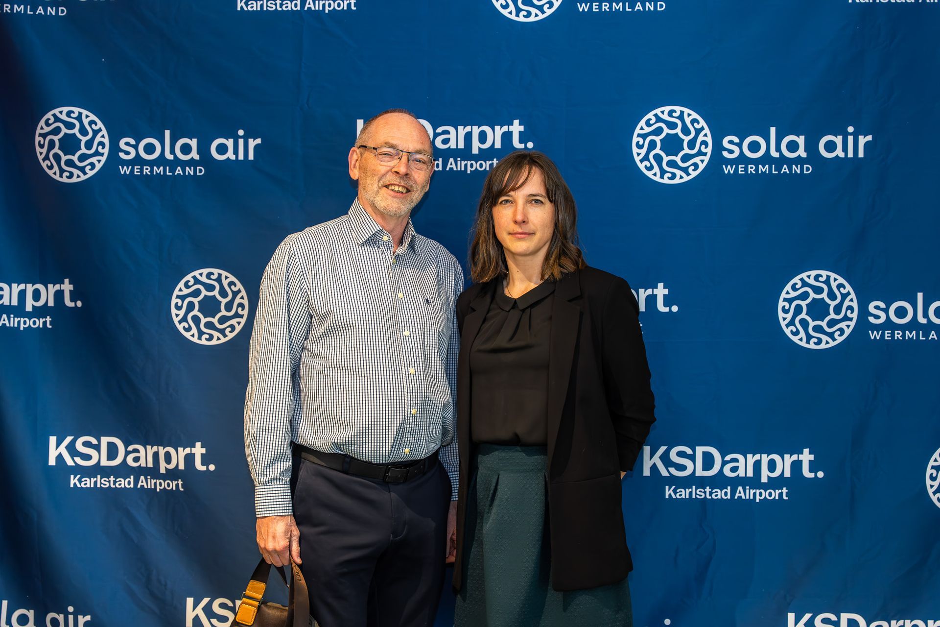 Två personer poserar i bakgrunden av ett KSDAirport/Sola Air-evenemang, iklädda affärskläder.