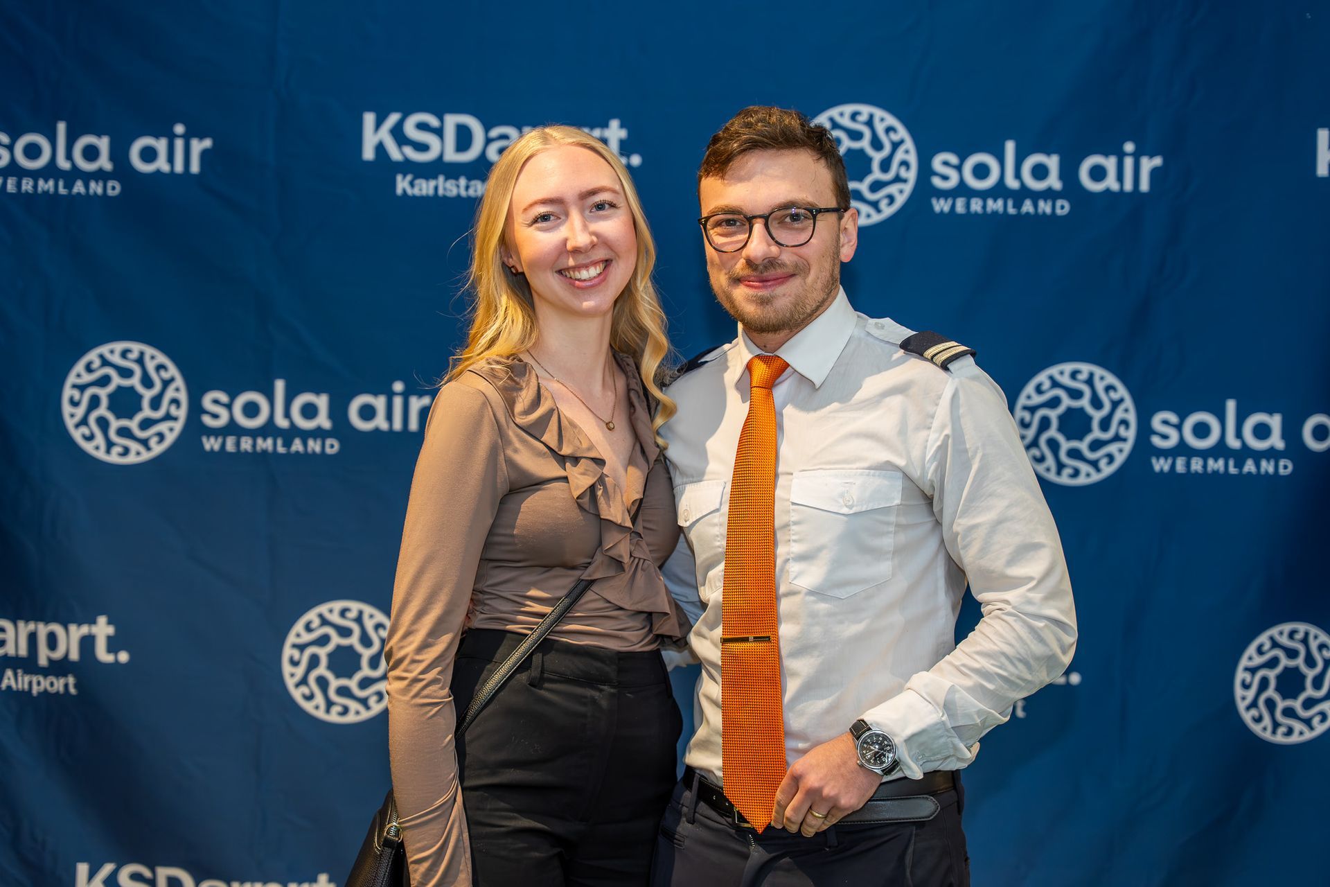 Två leende personer poserar framför en blå Sola Air- och Karpart-evenemangsbakgrund