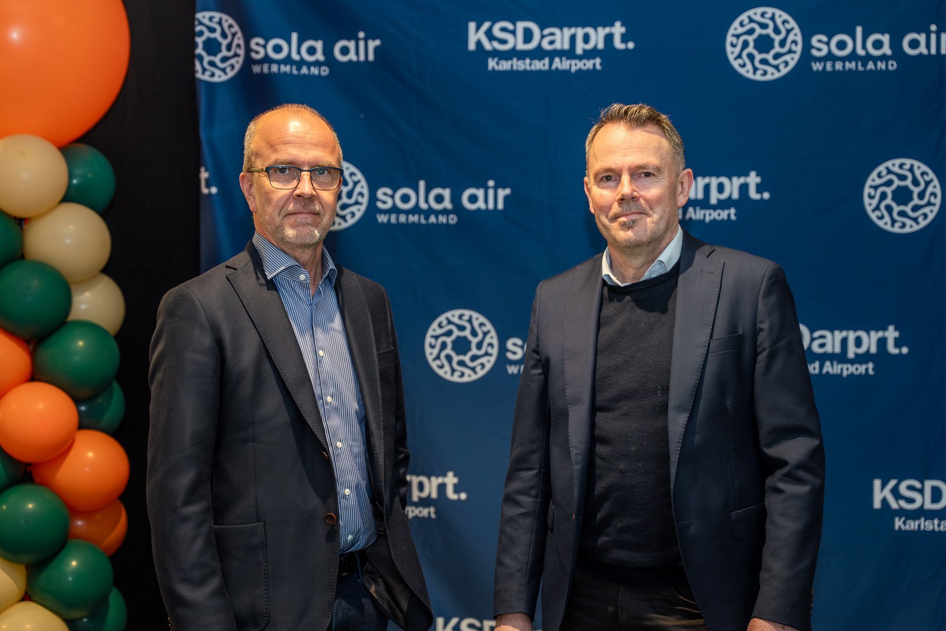 Två män i kostymer poserar i bakgrunden av ett evenemang för en flygplats i Kansas City och Sola Air, med orange ballonger till vänster.