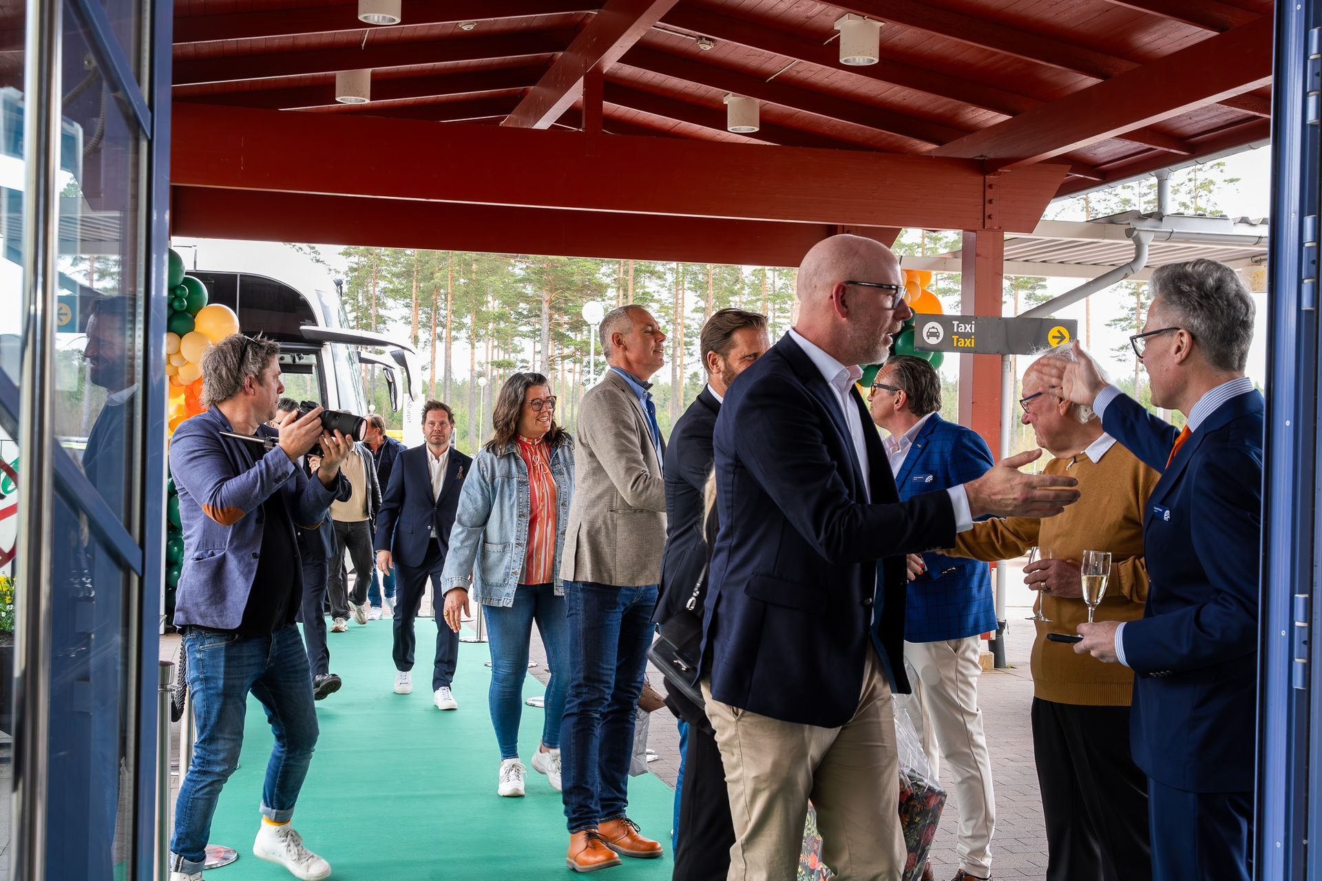 Människor minglar och hälsar på varandra under en gångväg med rött tak, med ett handslag i förgrunden.