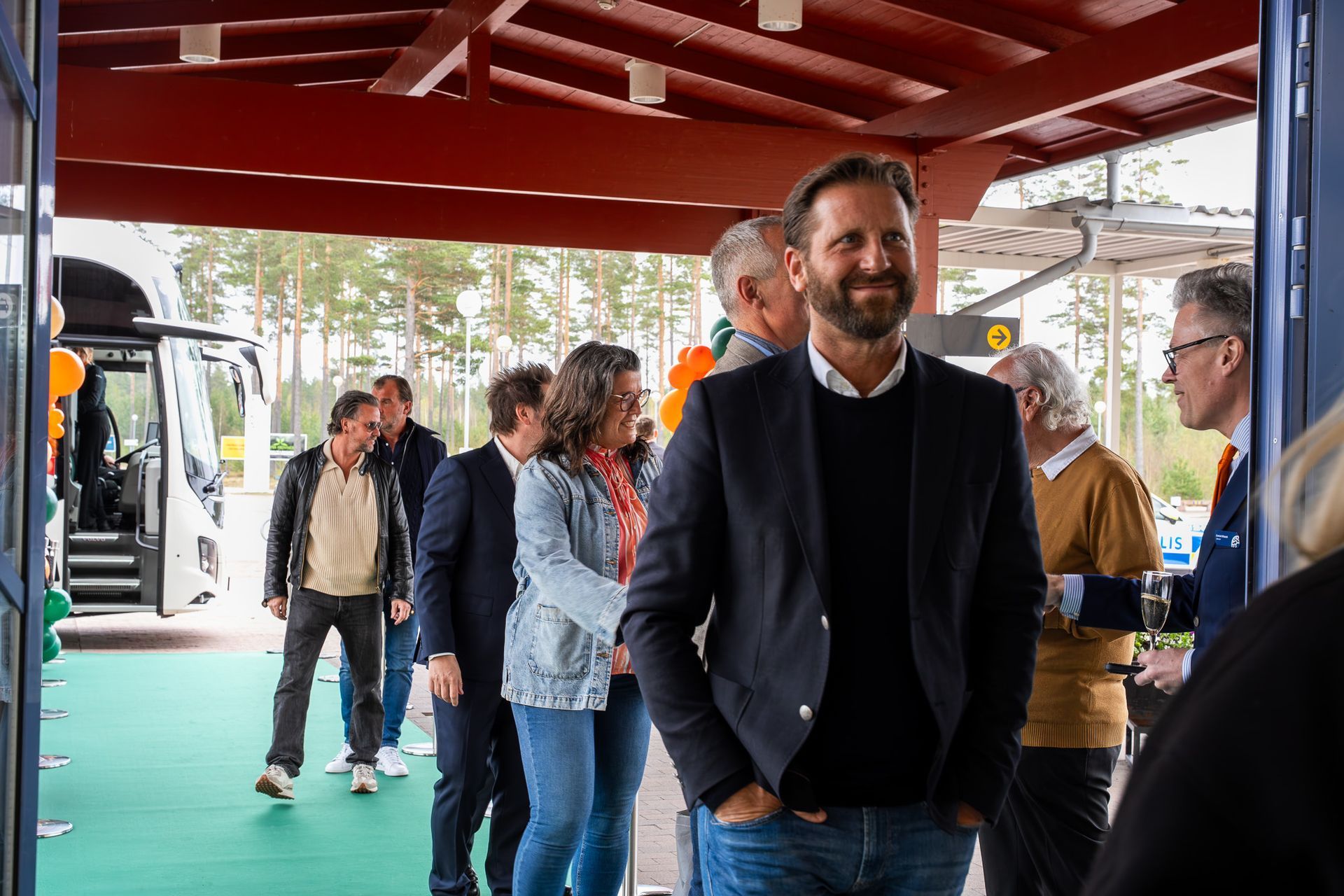 Människor som står under en utomhuspaviljong med rött tak, med en leende man i en marinblå tröja i förgrunden