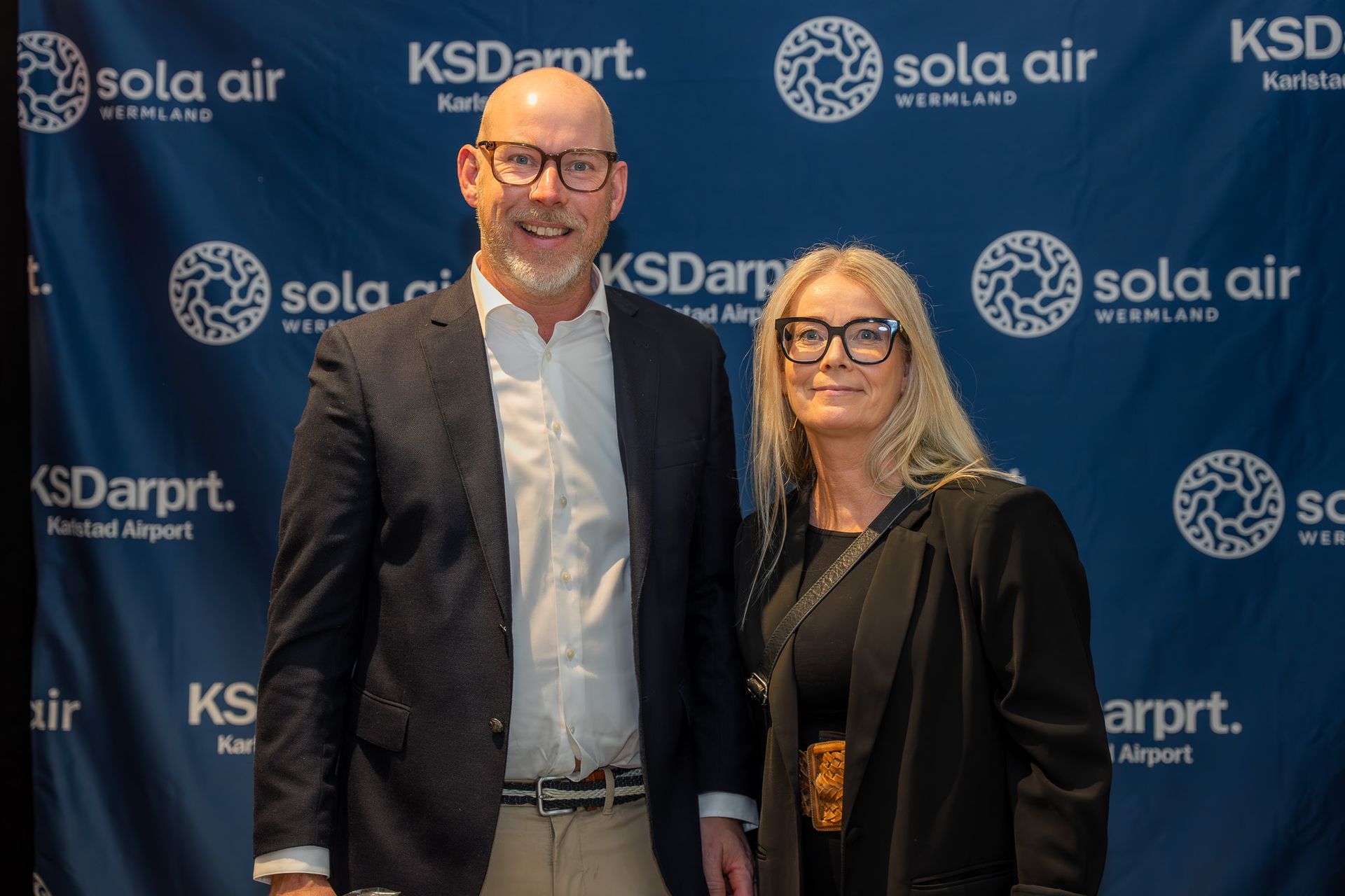 Två leende personer poserar framför en blå bakgrund av Sola Air och KSD flygplats
