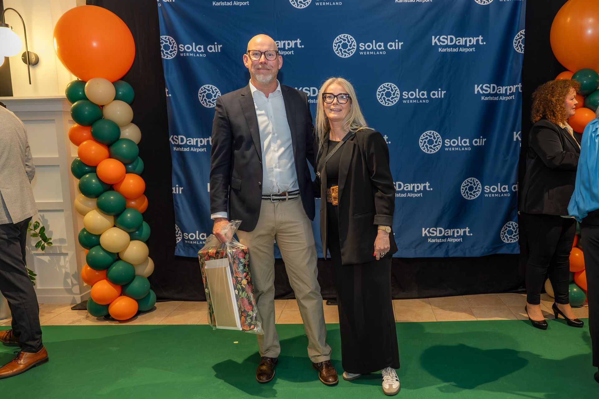 Två personer poserar vid en konferensbakgrund med ballonger och grön matta